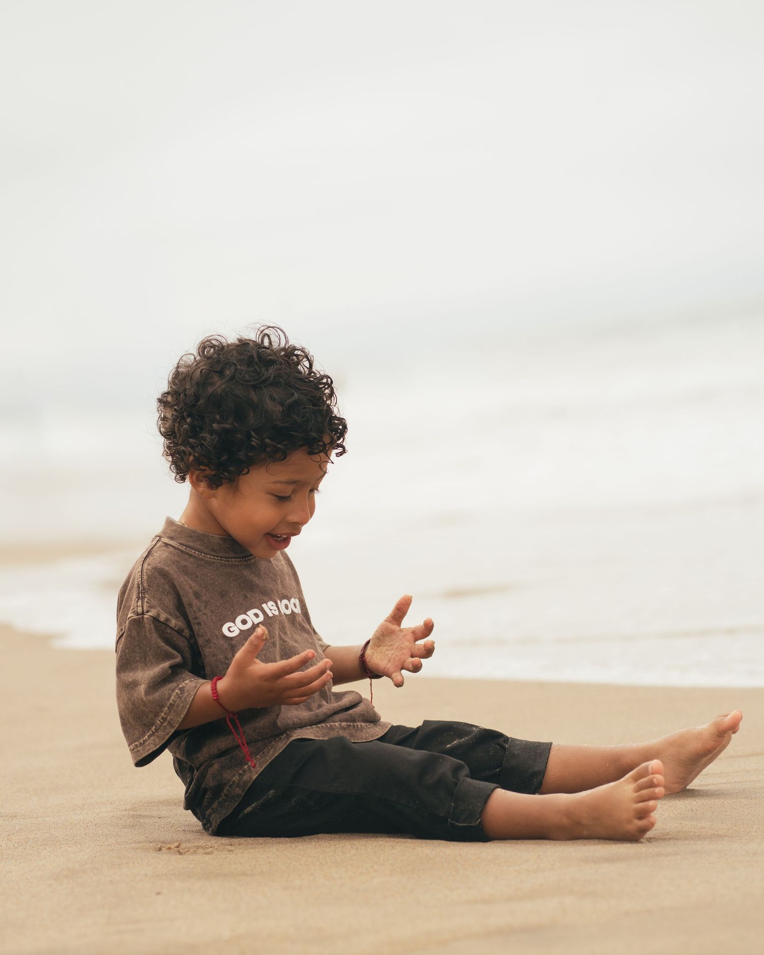 A curly-haired toddler sits barefoot on sandy beach, smiling while playing with sand near ocean waves.