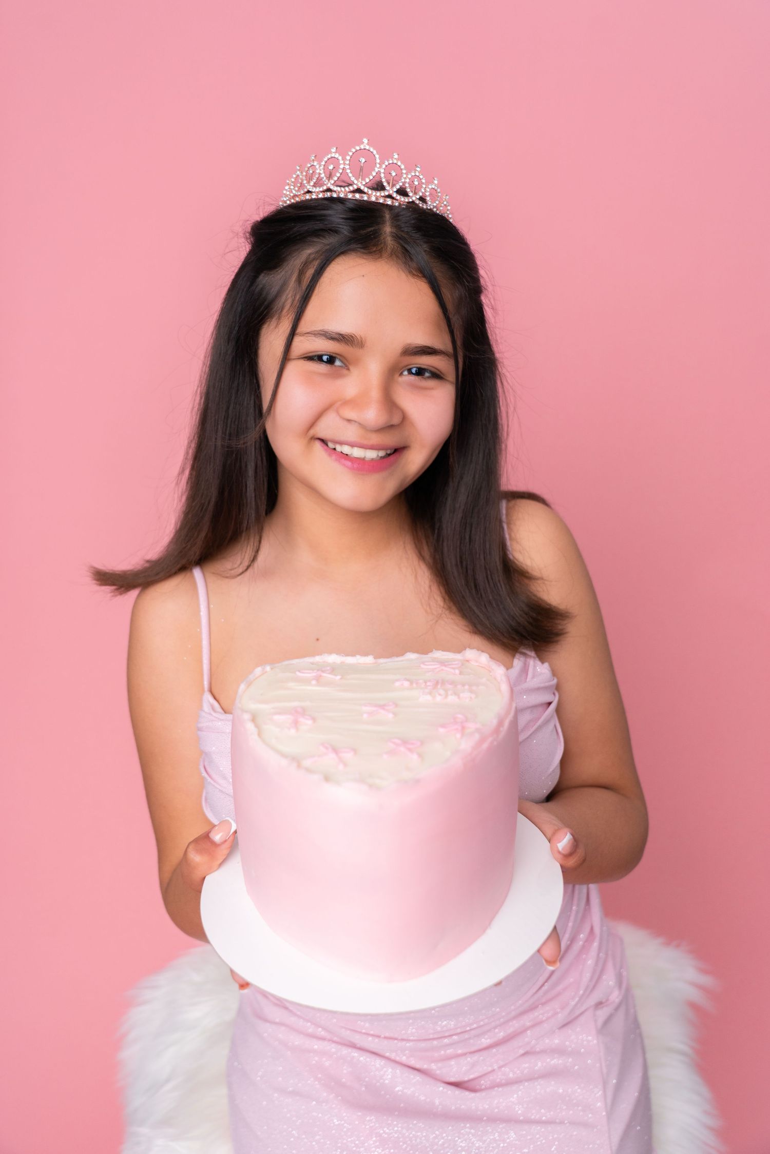 Child wearing pink dress and tiara poses with birthday cake against pink background.