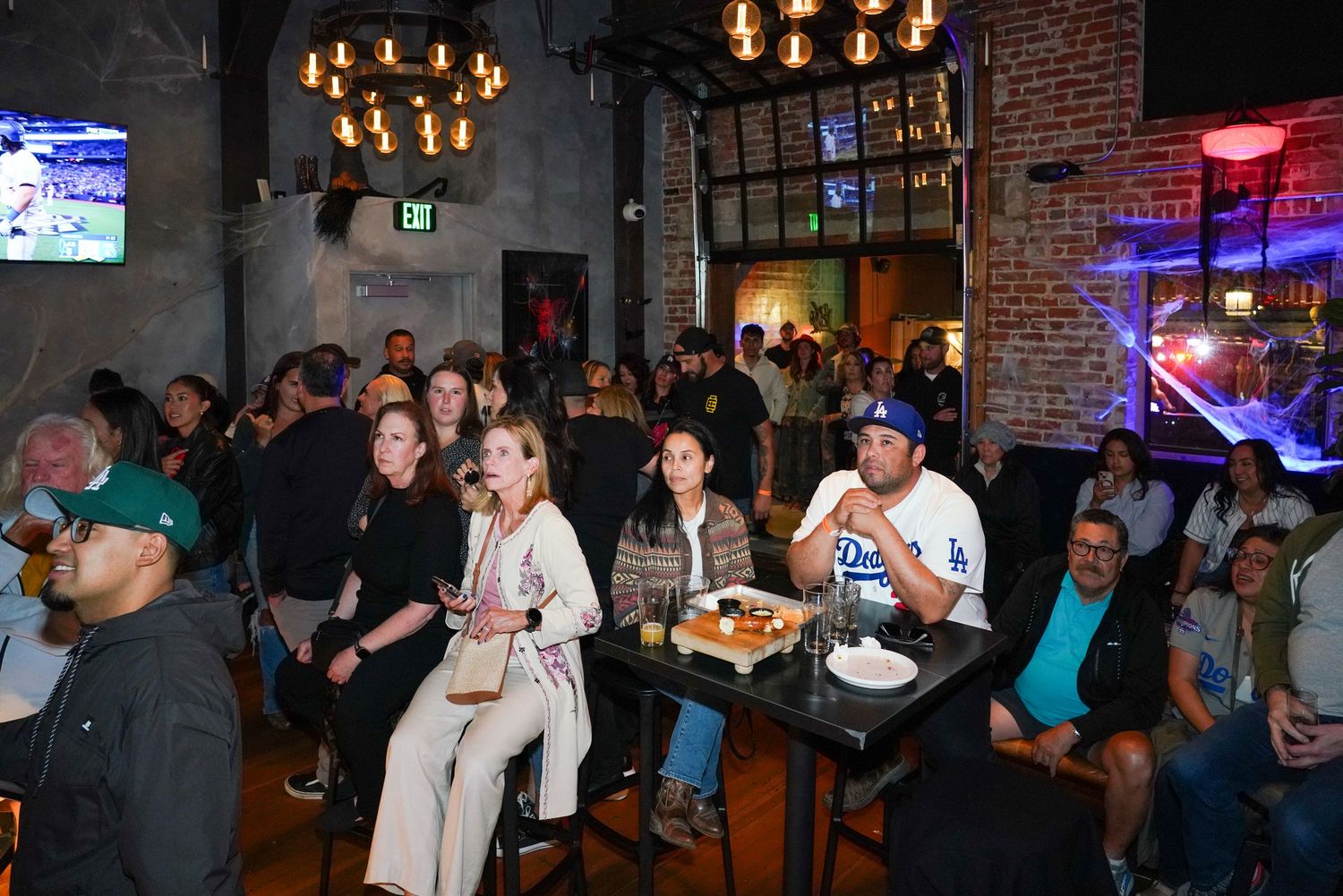 Crowd of sports fans watching a game on TV at a lively bar with brick walls and dim lighting.