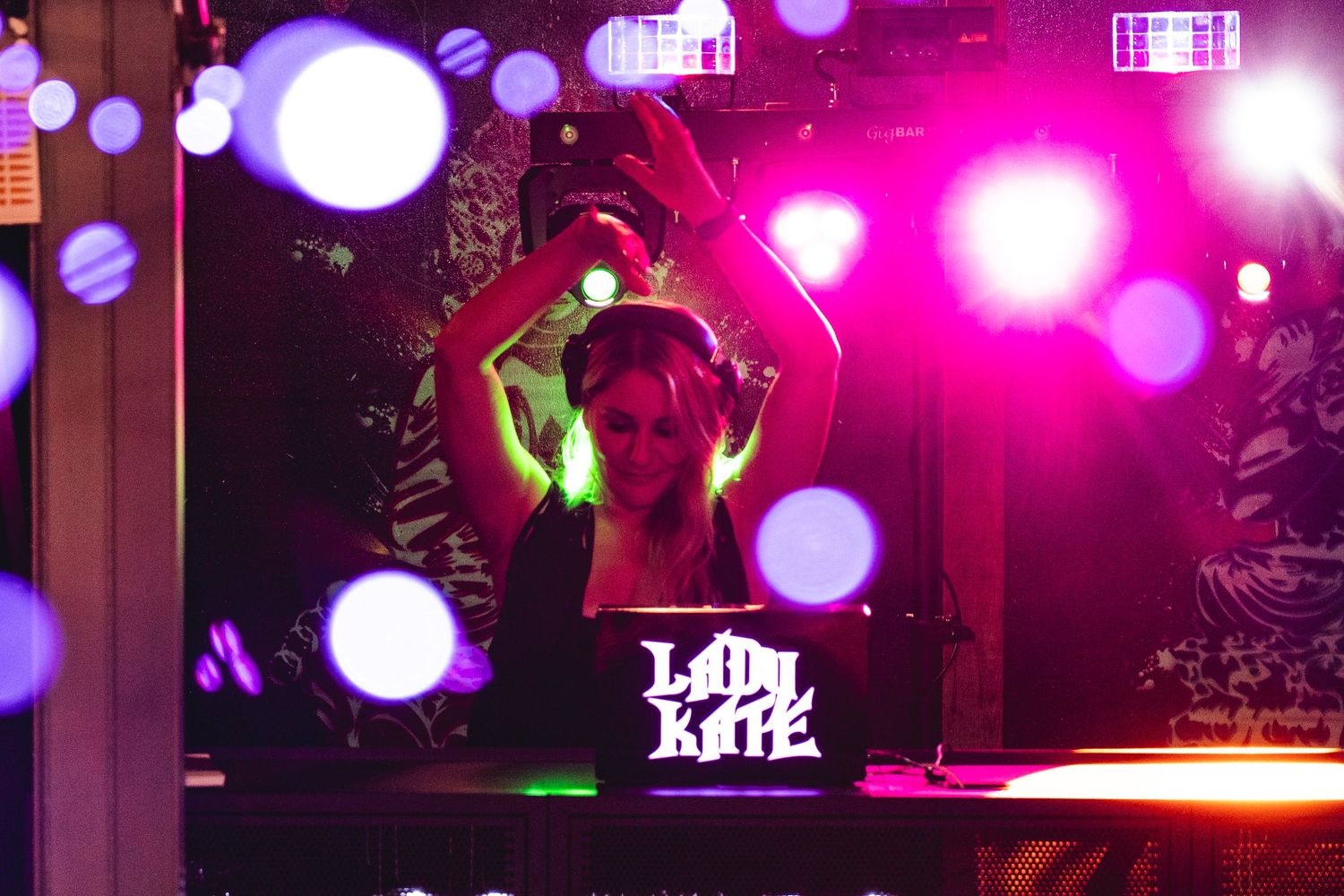 DJ Lady Kate performing under colorful stage lights with hands raised behind a glowing DJ booth at a nightclub event.