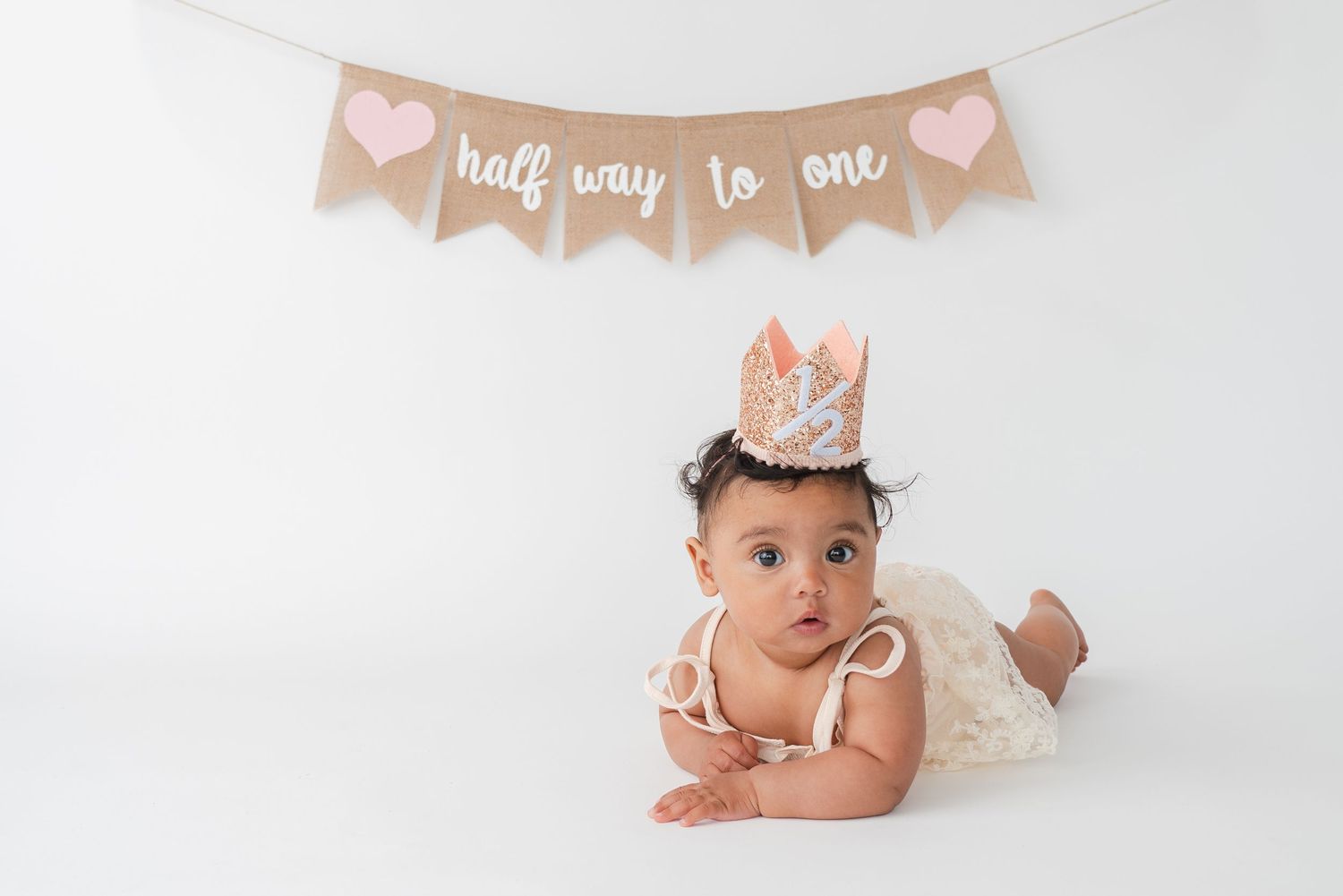 Baby wearing pink birthday crown lays on white background with half birthday decorations and milestones.