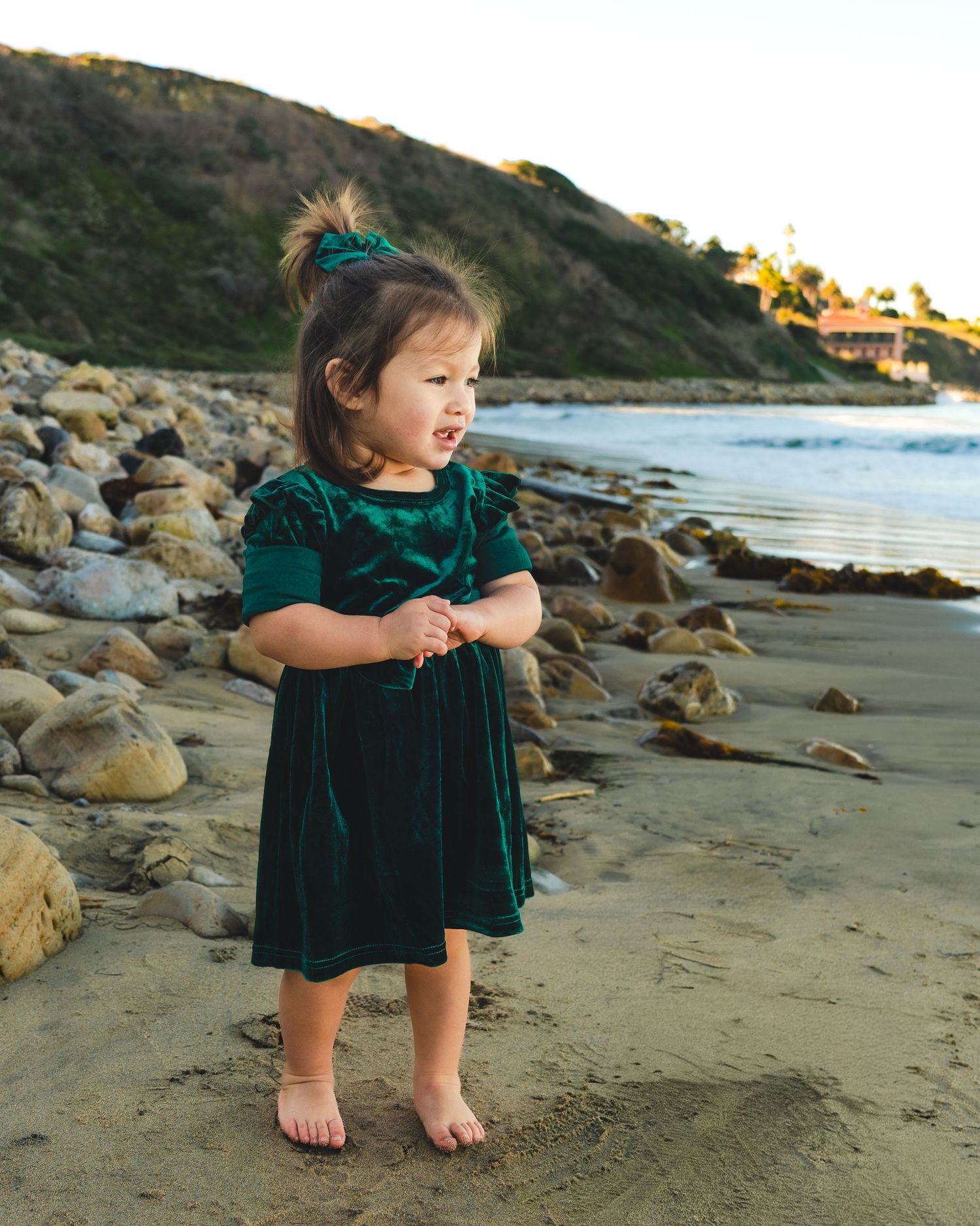 A child wearing an emerald velvet dress sits thoughtfully on a rocky beach at sunset.