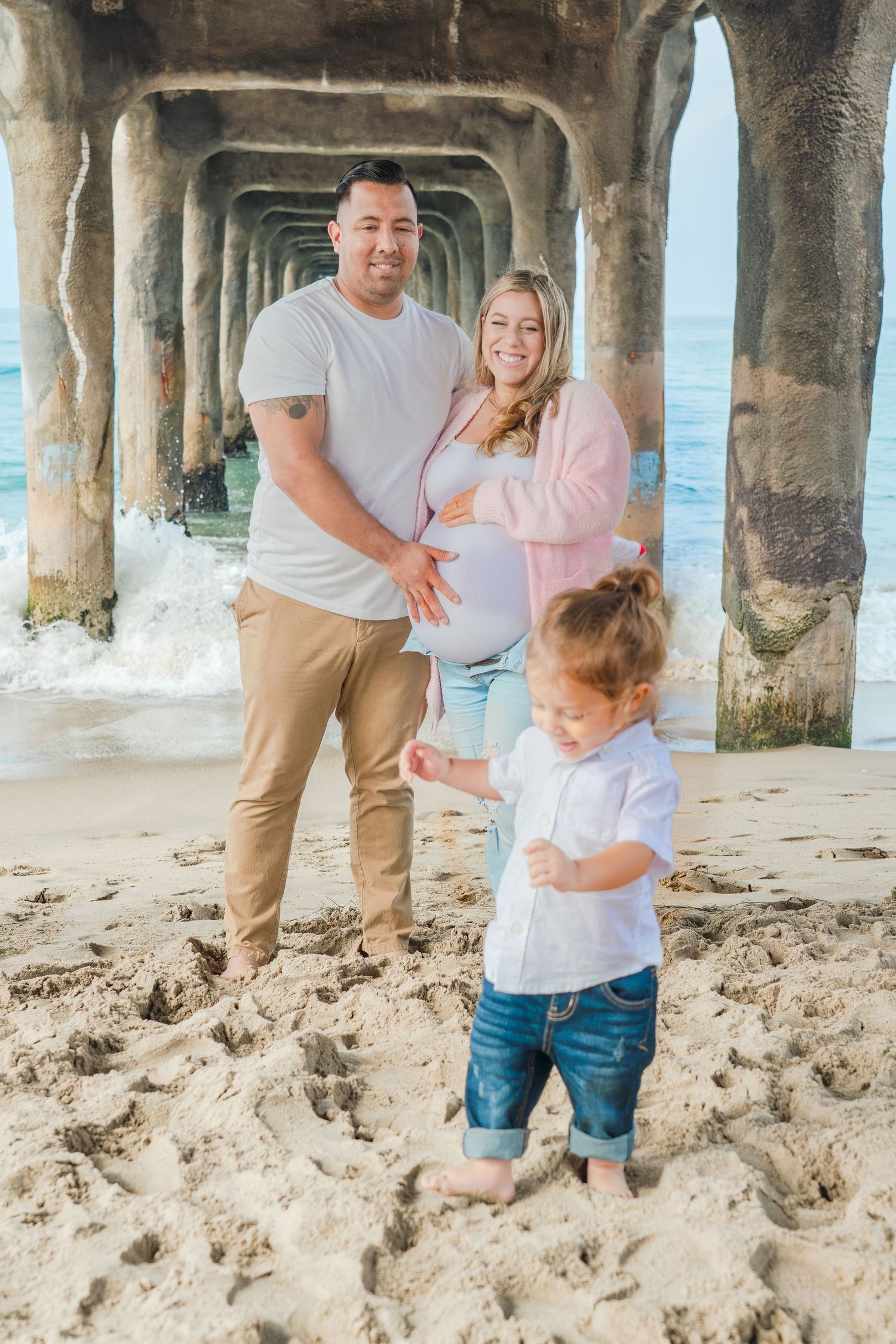 Family enjoying playful moments under a pier at the beach wearing light casual clothes.