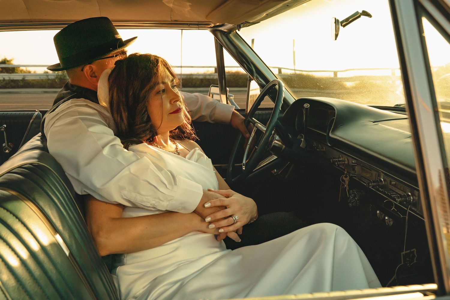 Couple embracing in a vintage car at sunset, woman in white dress and man in hat, romantic golden hour photography.