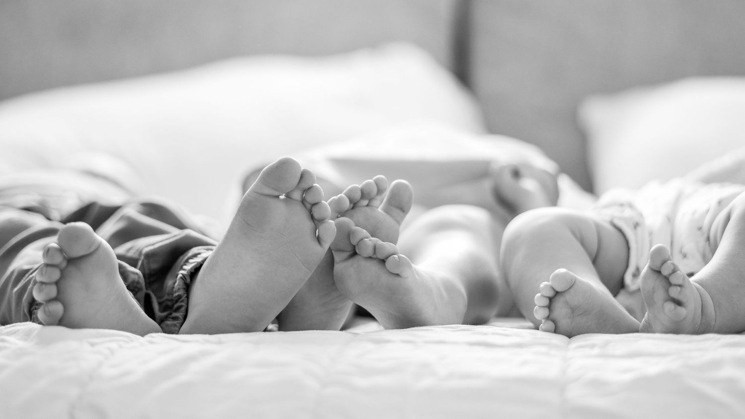 Black and white photo of adult and baby feet resting together on a soft bed, capturing a tender family moment.