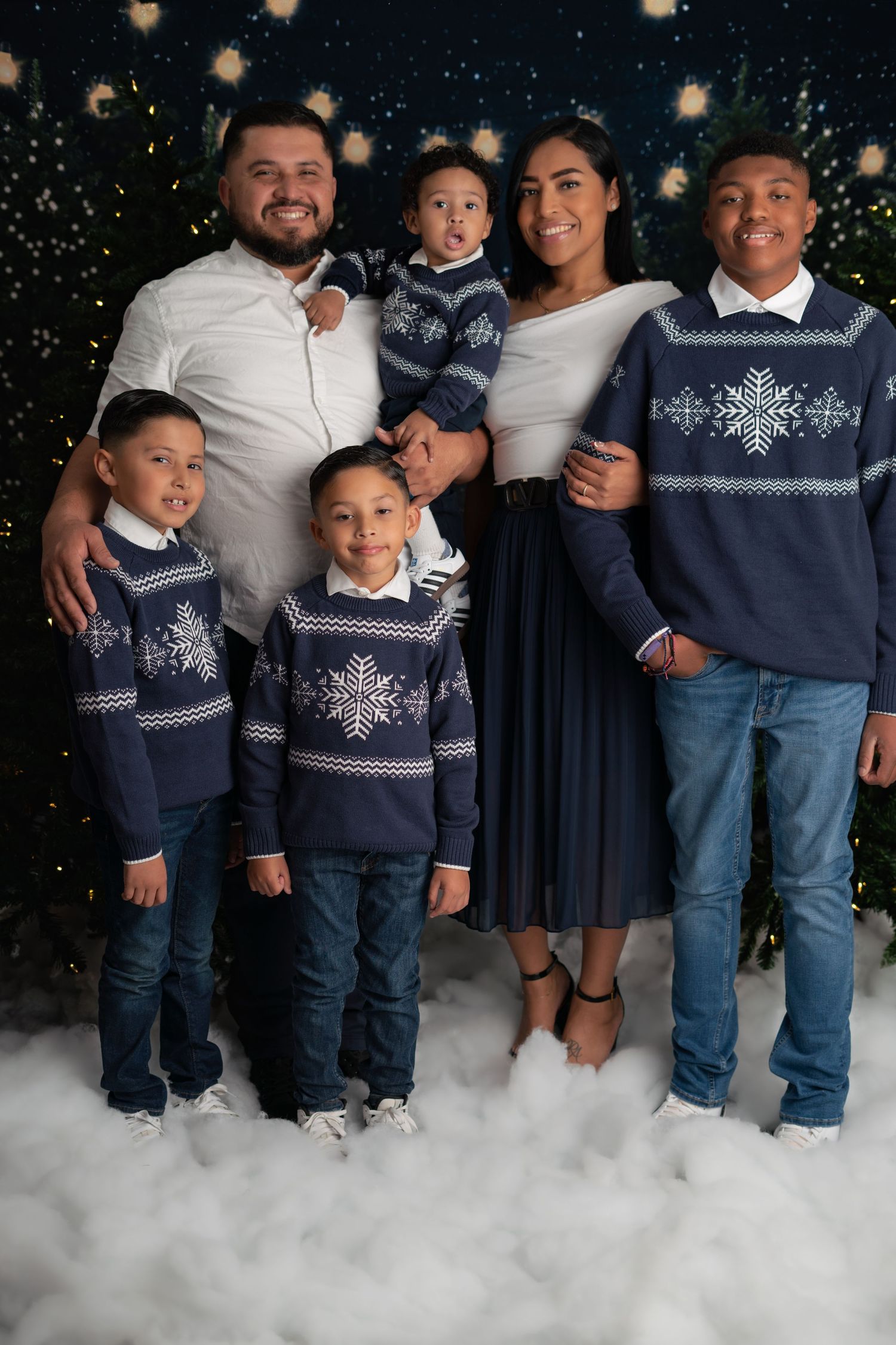 Family wearing coordinated navy and white winter outfits pose together in studio with artificial snow effect.