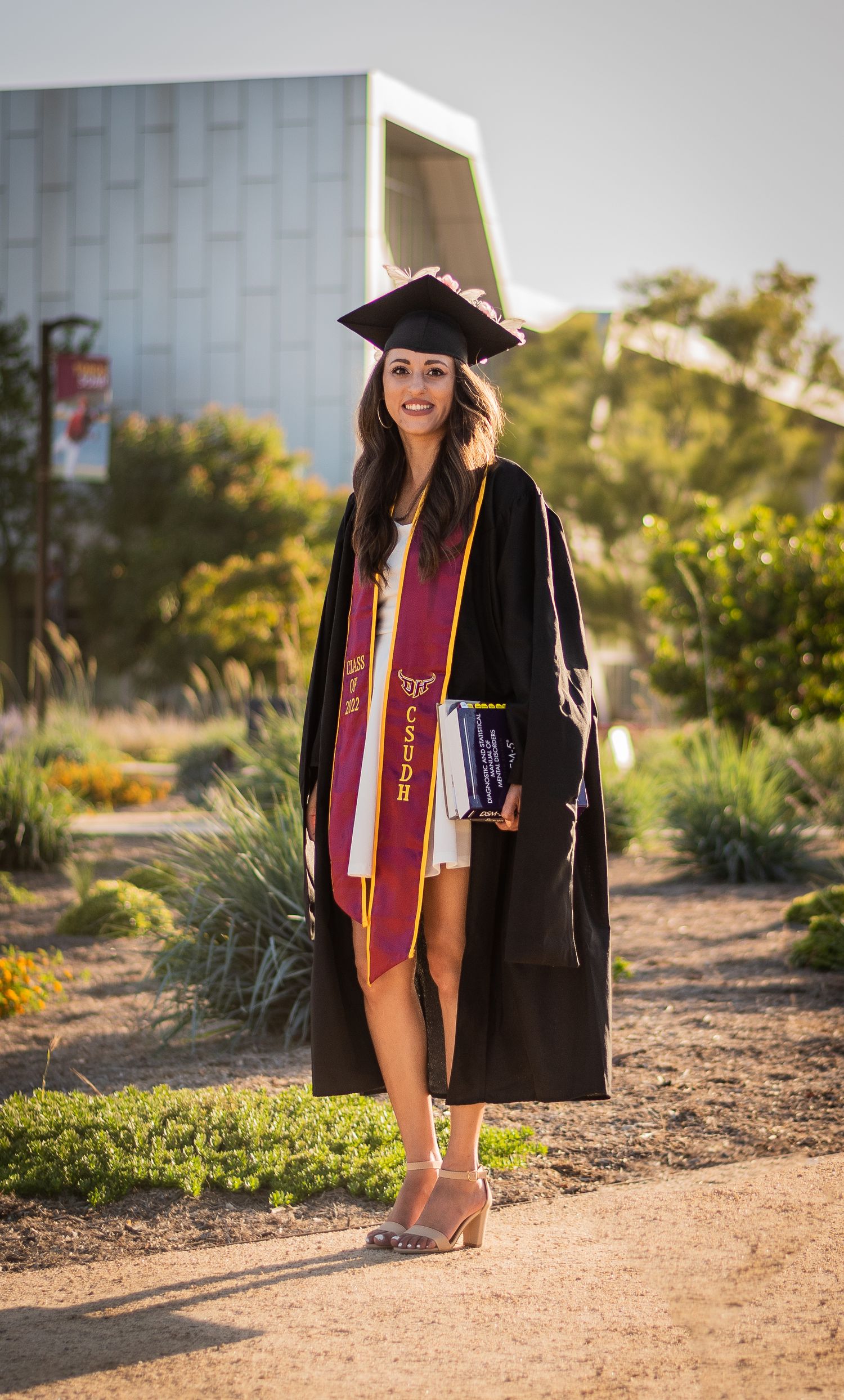 Graduate in cap and gown with maroon sash poses outdoors on campus path during golden hour celebration.