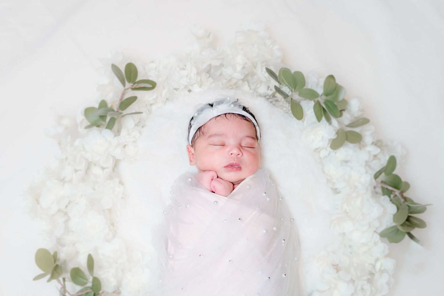 Newborn photography setup with white flowers and green leaves arranged in circle.