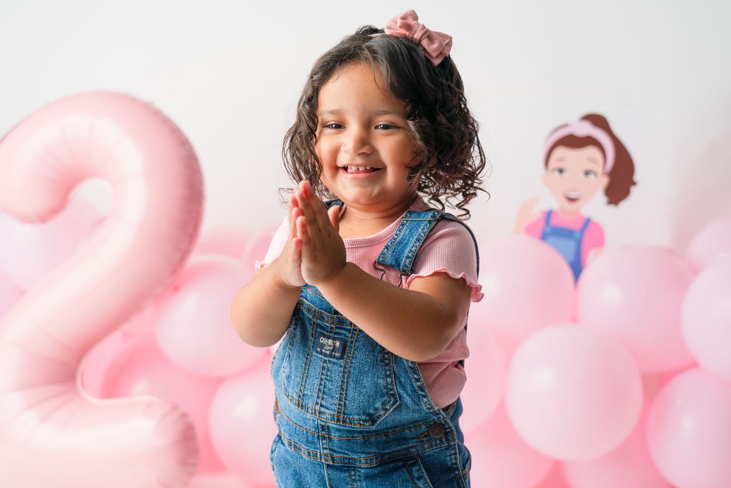 Child in overalls poses playfully with pink balloons in studio setting.