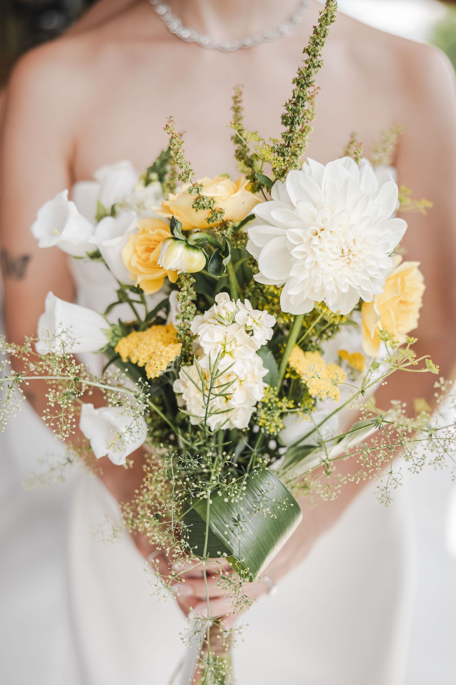 Delicate yellow and white wedding bouquet with wildflowers and greenery held against a light background.