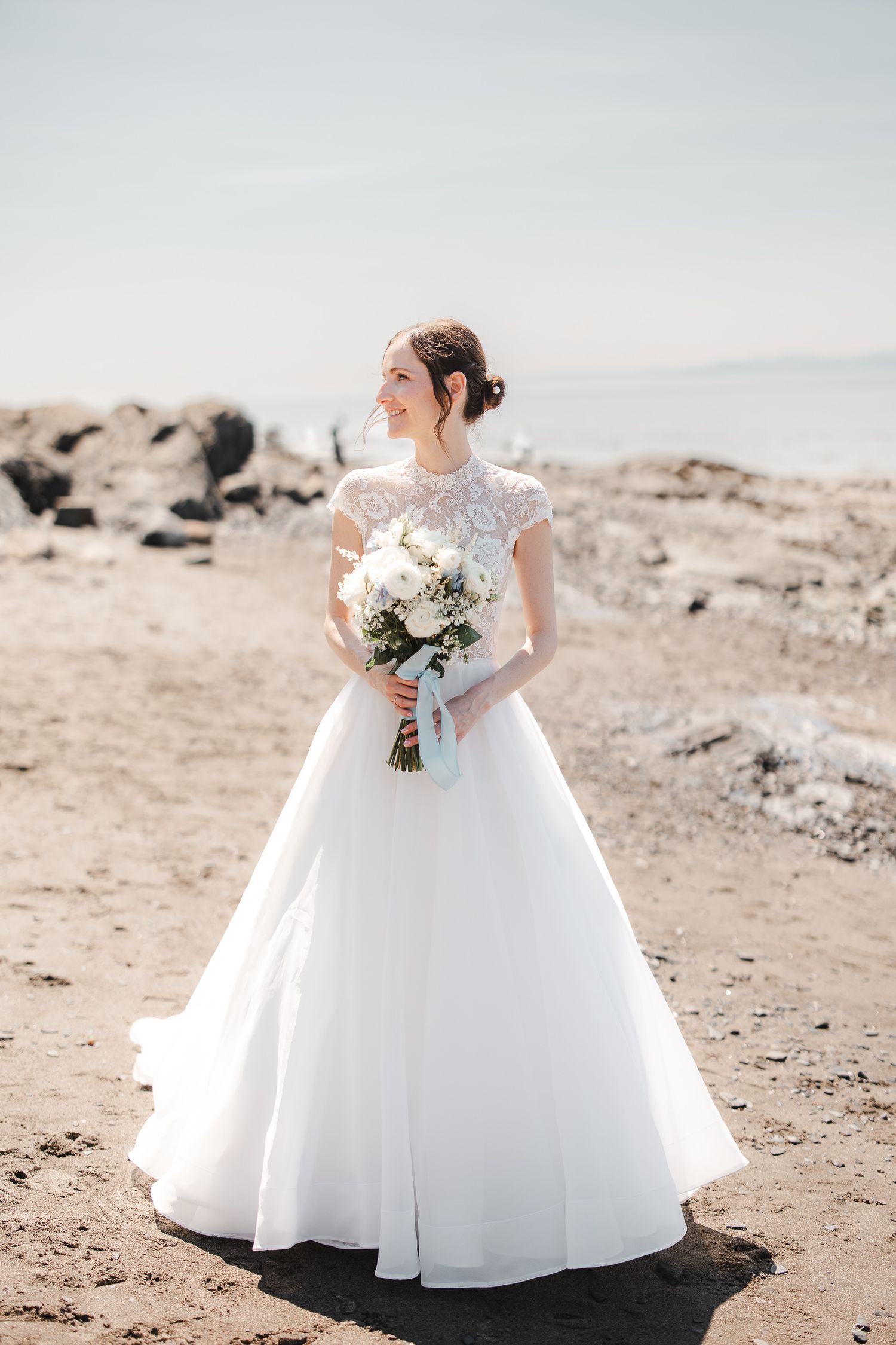 Mariée en robe de bal blanche tenant un bouquet floral blanc, photographiée sur une plage rocheuse lors de son mariage.