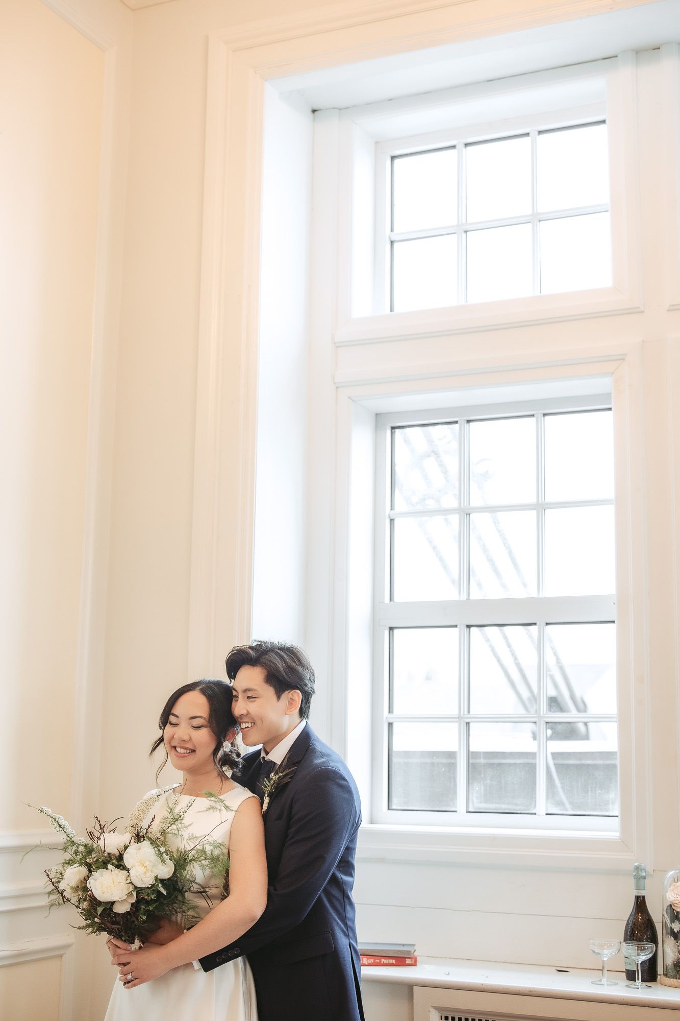 Moment de first look intime d’un couple dans une pièce blanche lumineuse avec grandes fenêtres et bouquet floral