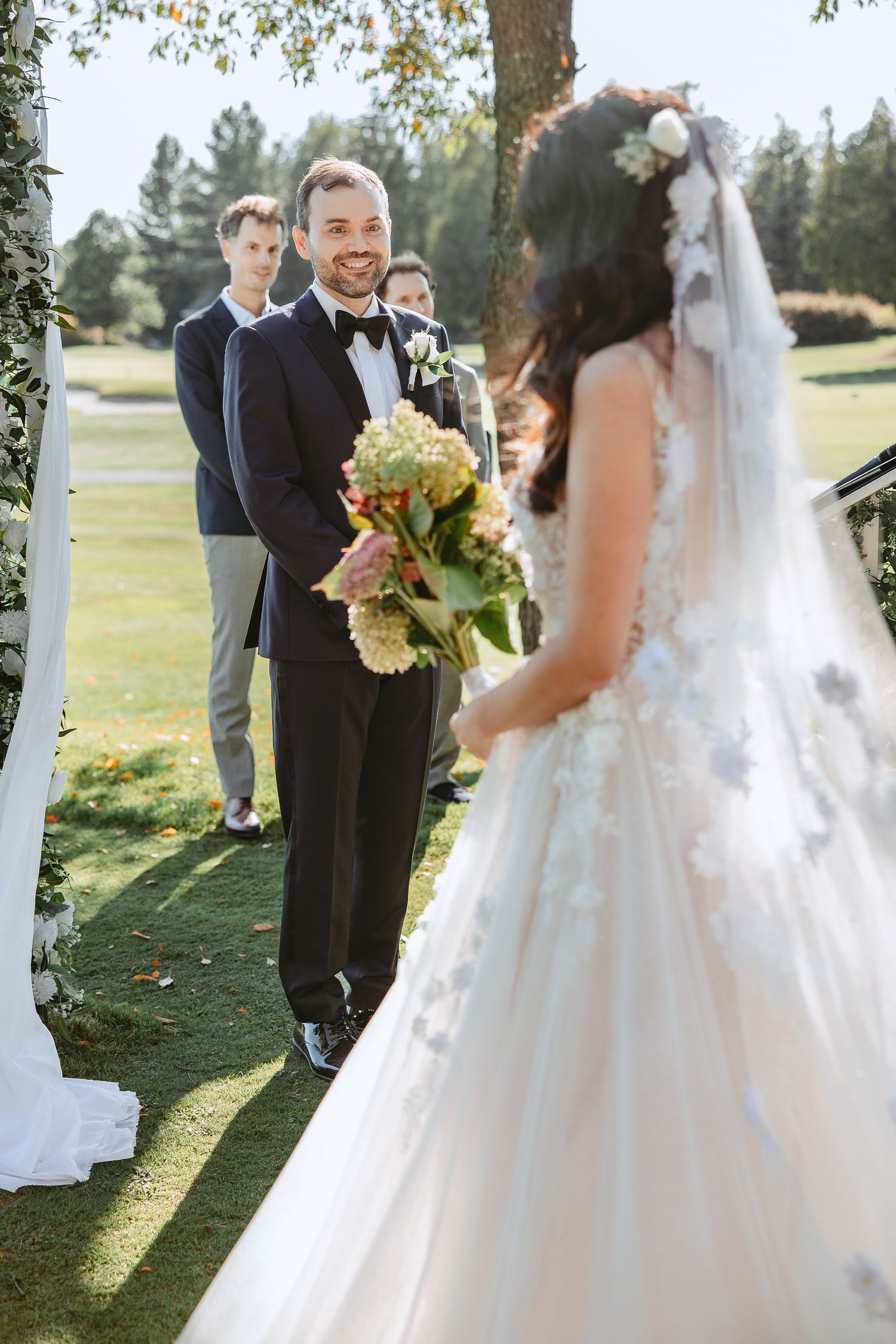 Wedding ceremony taking place outdoors on a sunny day with white decor and floral arrangements on a grassy lawn.