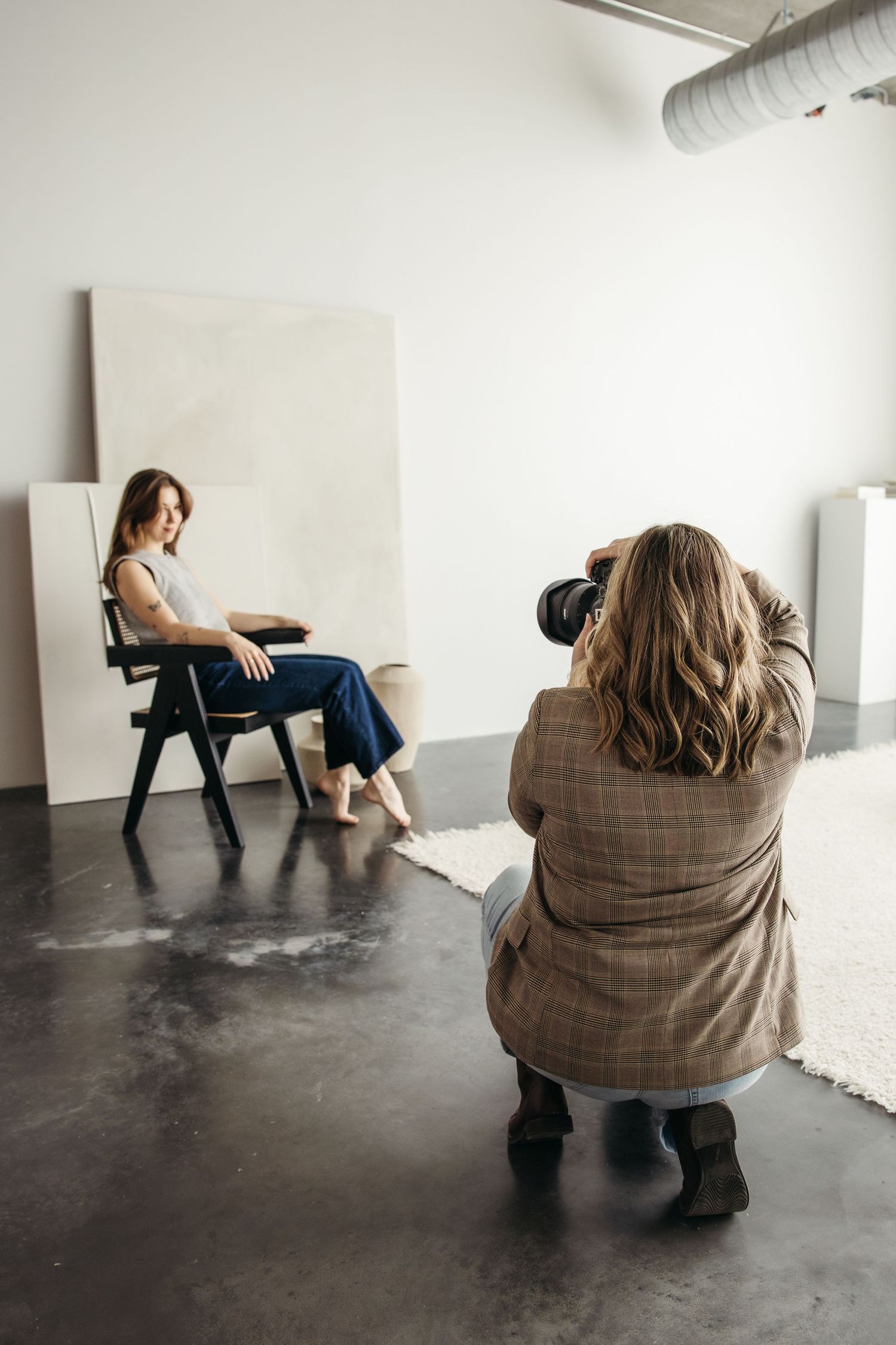 A photographer in a brown blazer crouches to take photos of a model seated in front of a white backdrop in a studio.