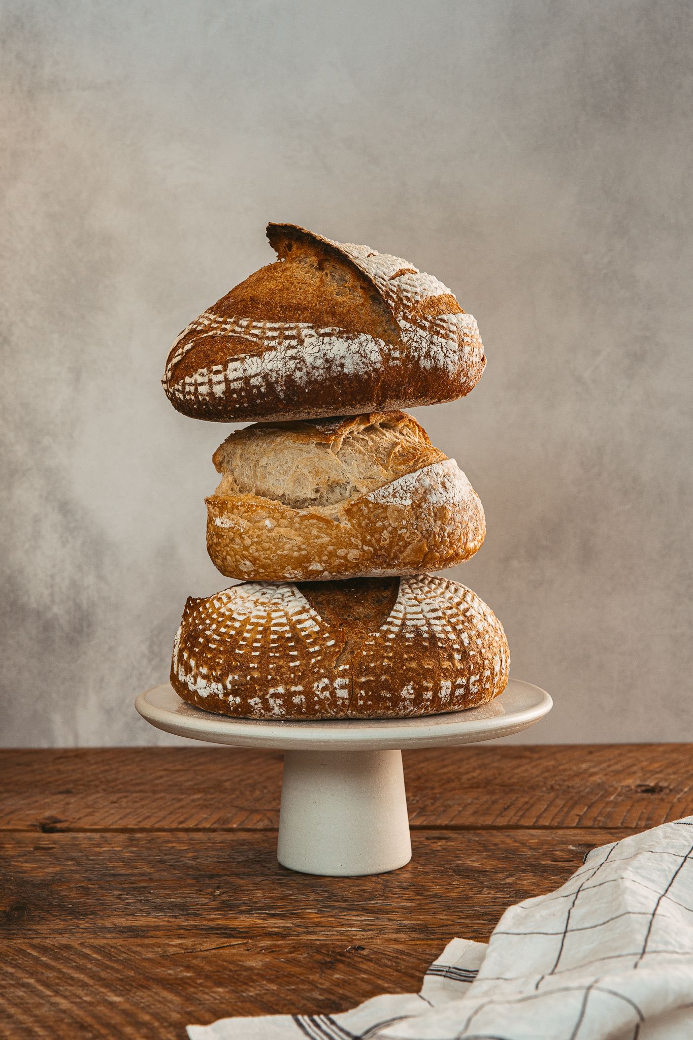 Artisanal bread loaves stacked on white cake stand against textured backdrop.