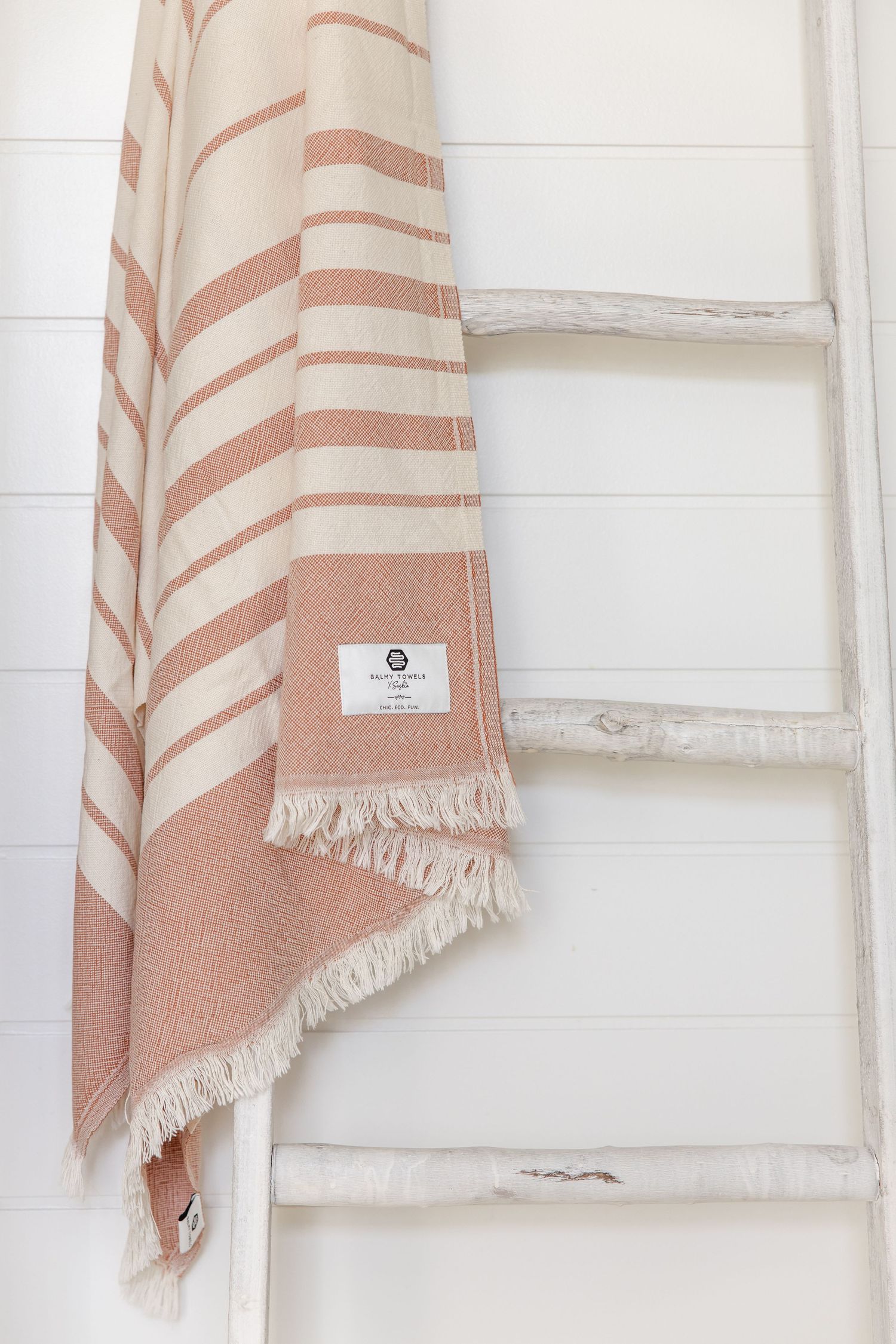 A soft beige and white striped Turkish cotton towel with fringe tassels hangs against a white wall.