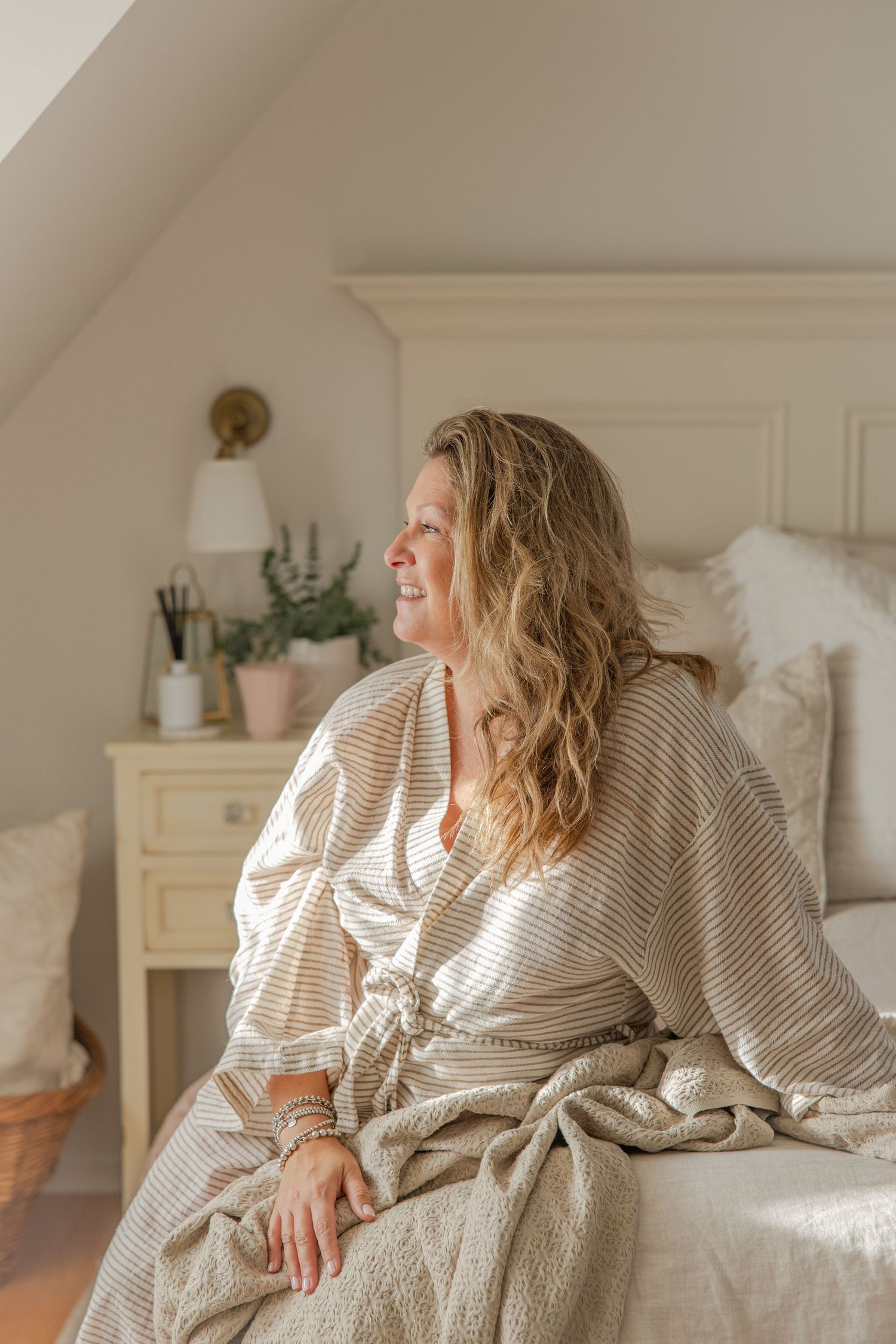 Person relaxing in cozy white bedroom wearing striped robe while natural light streams through window.