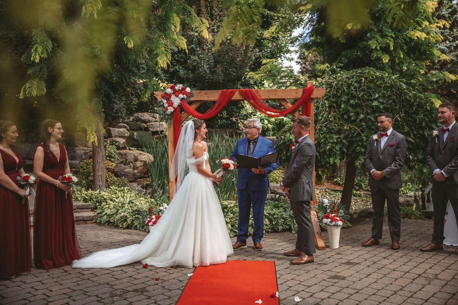 Outdoor garden wedding ceremony with guests seated on chairs along a stone pathway surrounded by lush greenery.