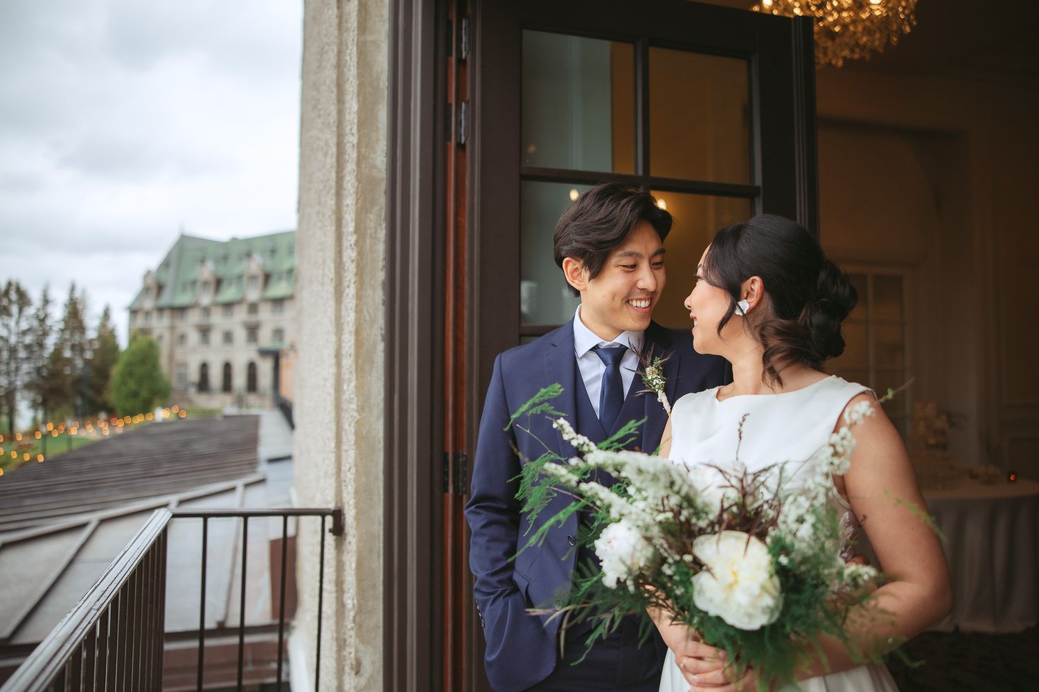 A romantic wedding couple shares an intimate moment on a balcony with elegant architecture and lighting.