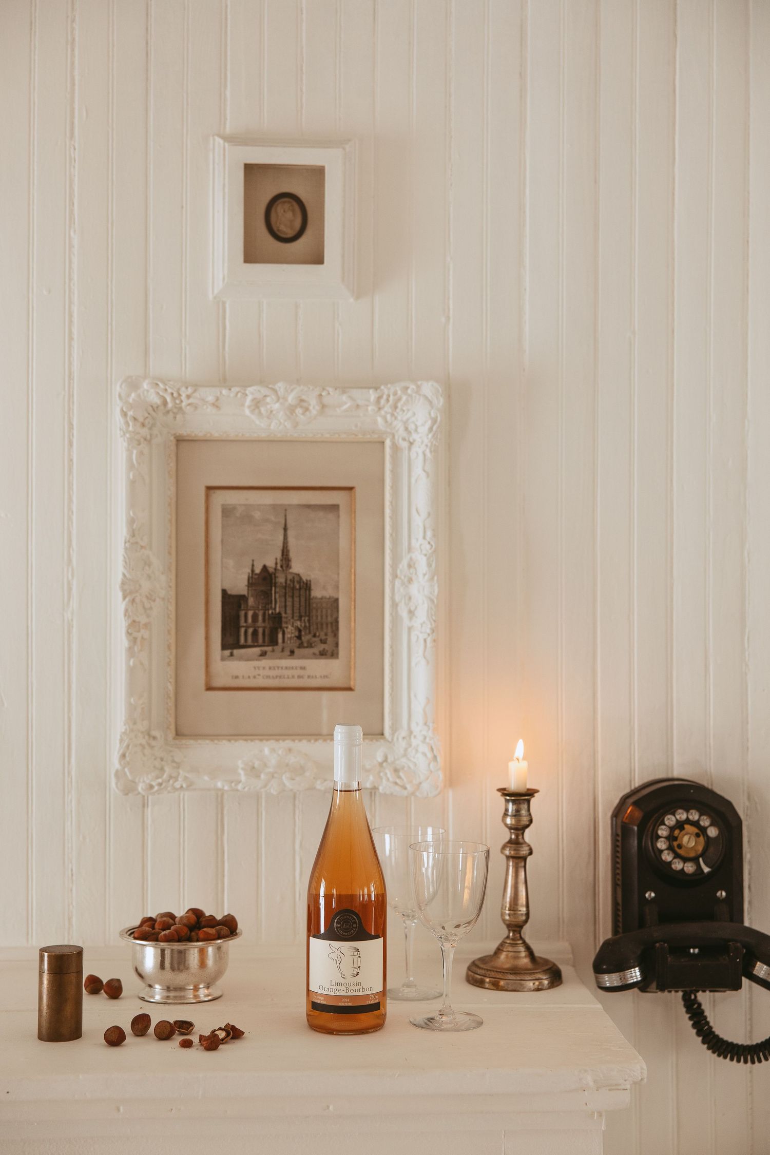 Vintage decor arrangement on white shelf with candlelight, old telephone, wine bottle, and framed artwork.