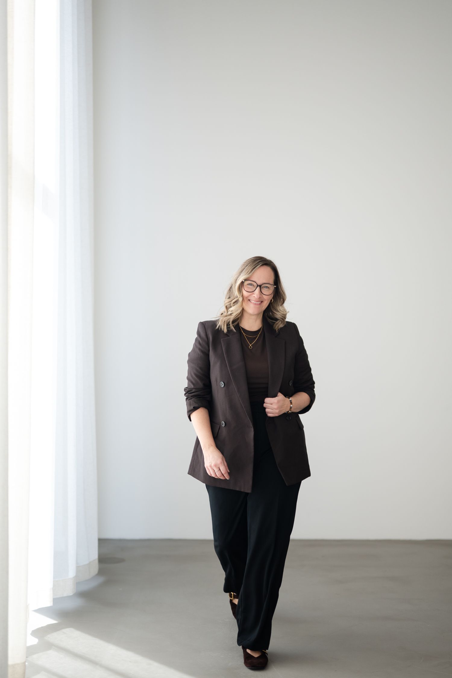 Professional in black attire poses in a minimalist white studio space with natural lighting from windows.