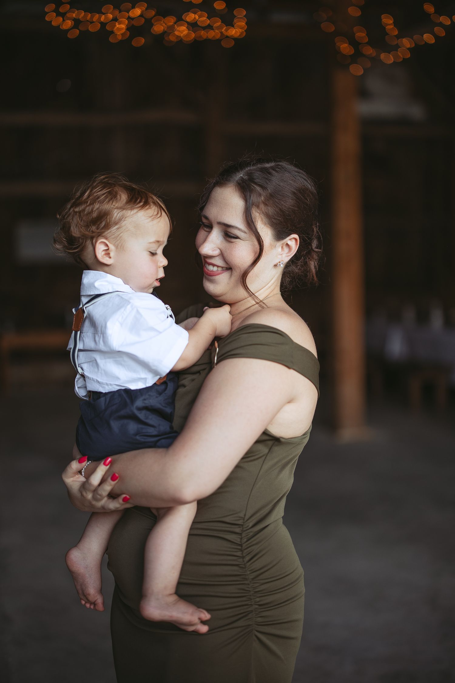 Mère en robe vert olive tenant son tout-petit en chemise blanche et pantalon bleu marine, sur fond sombre