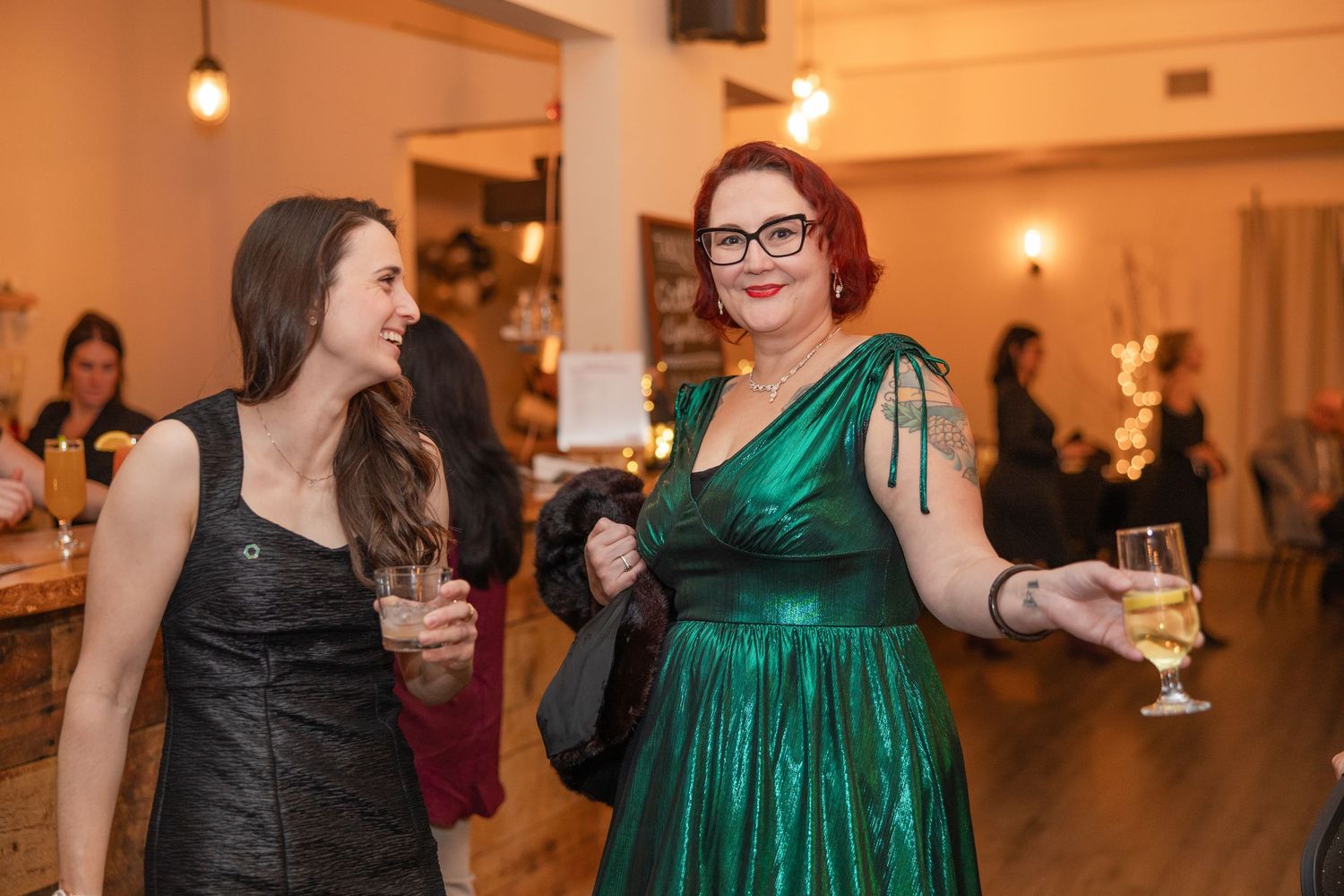 Two guests in formal attire share a laugh at an indoor event, one wearing black and one in a sparkly emerald dress.