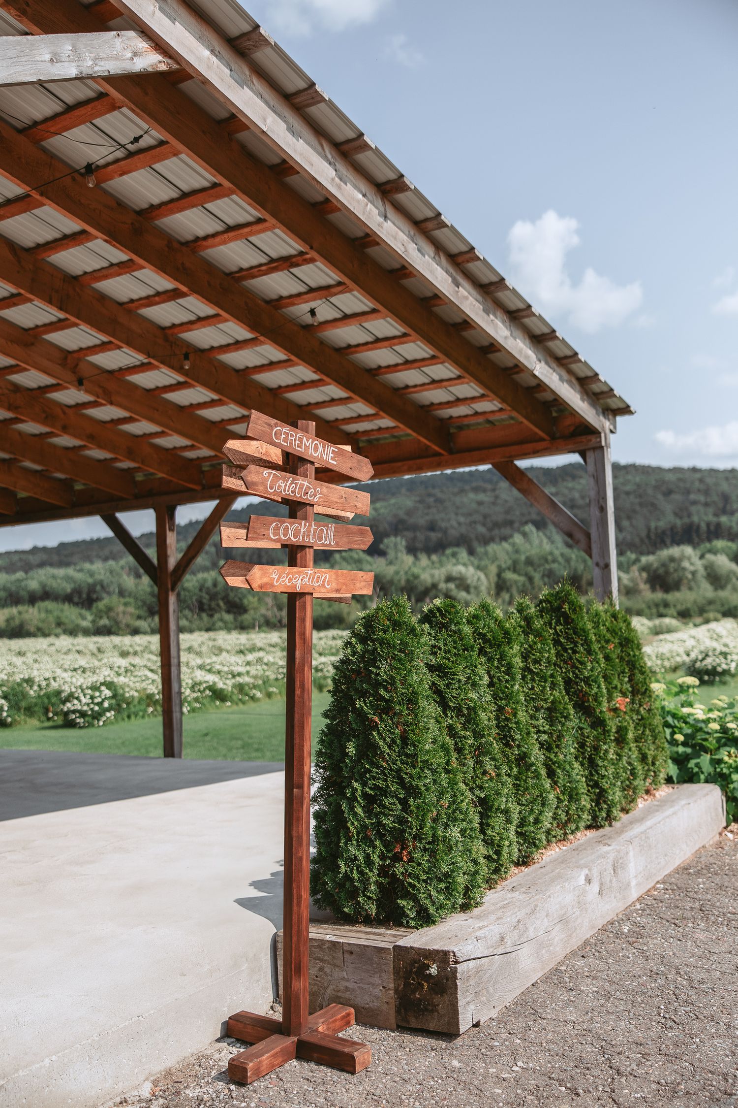 Panneau directionnel en bois sous un patio extérieur couvert, avec arbustes persistants et vue sur les montagnes en arrière-plan.