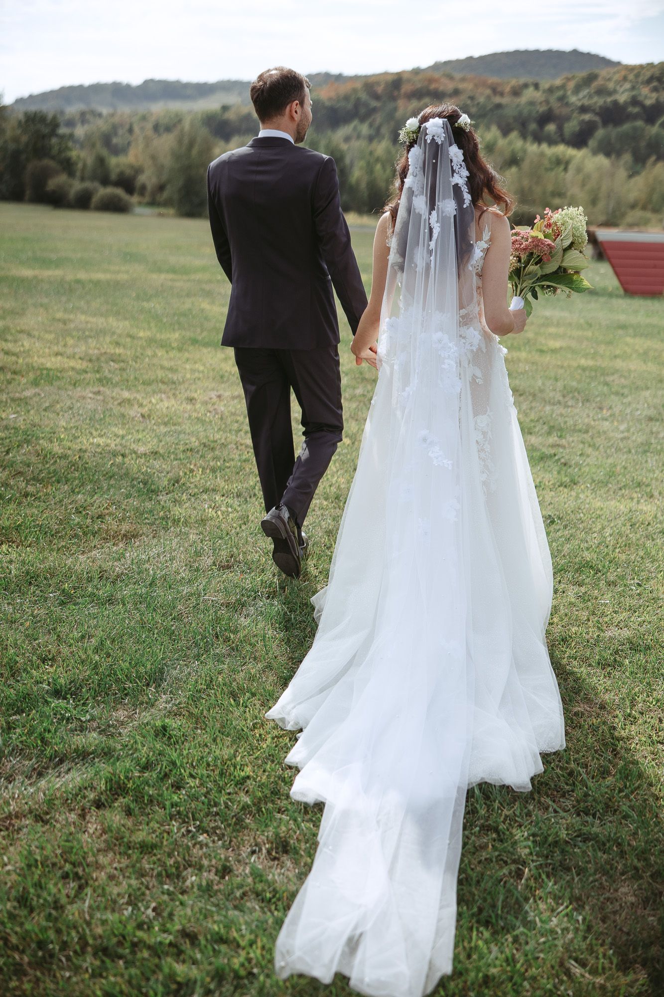 Mariés marchant ensemble sur une colline herbeuse le jour de leur mariage, la robe blanche flottant derrière la mariée