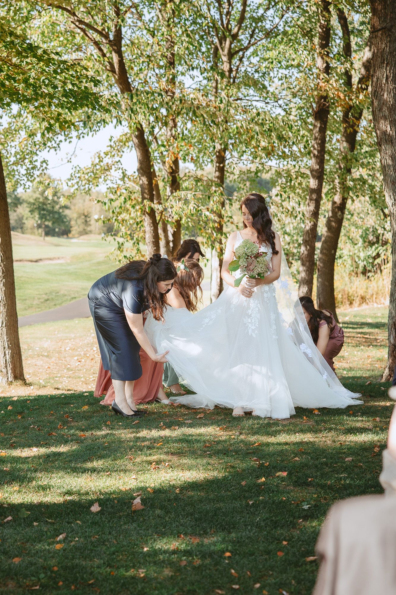 Demoiselles d’honneur aidant à ajuster la robe de mariée blanche fluide lors d’une journée d’automne ensoleillée, sous les arbres