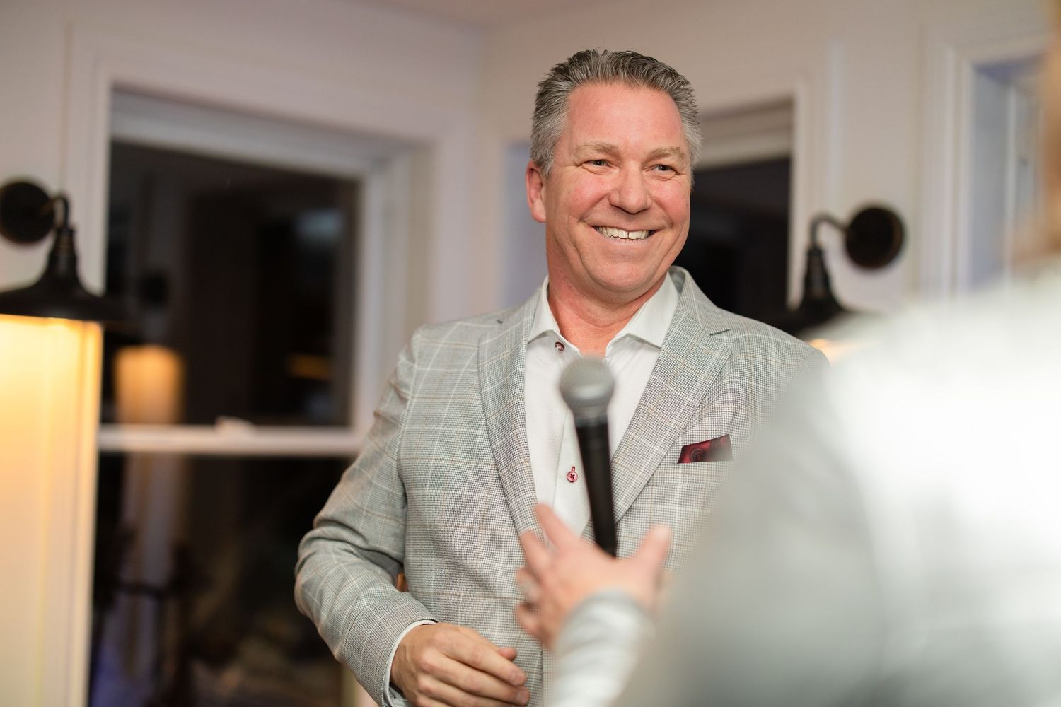 A person in a light colored suit smiles warmly while standing in a well-lit indoor space.