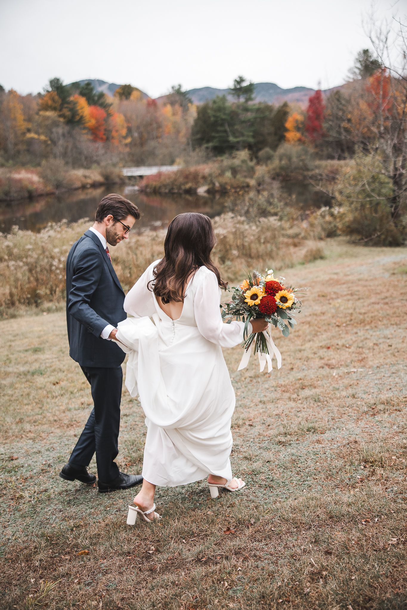 Un couple en tenue de mariage marche ensemble sur une colline herbeuse en automne, avec un feuillage coloré en arrière-plan.