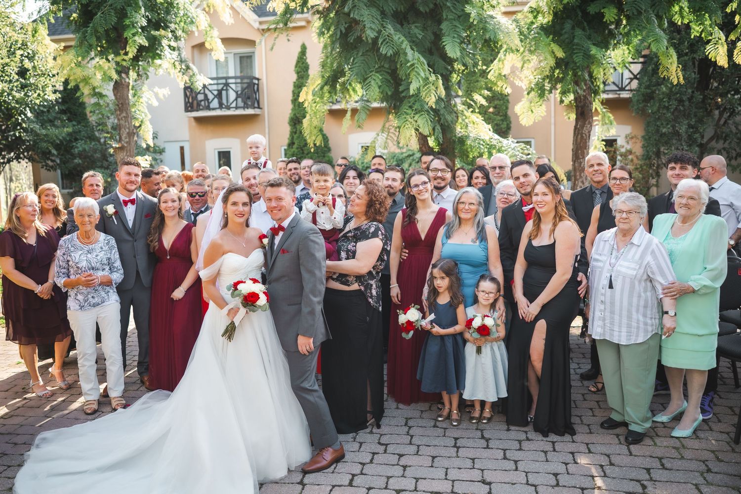 Large wedding party gathered outdoors on brick patio with guests wearing formal attire in burgundy and neutral colors.