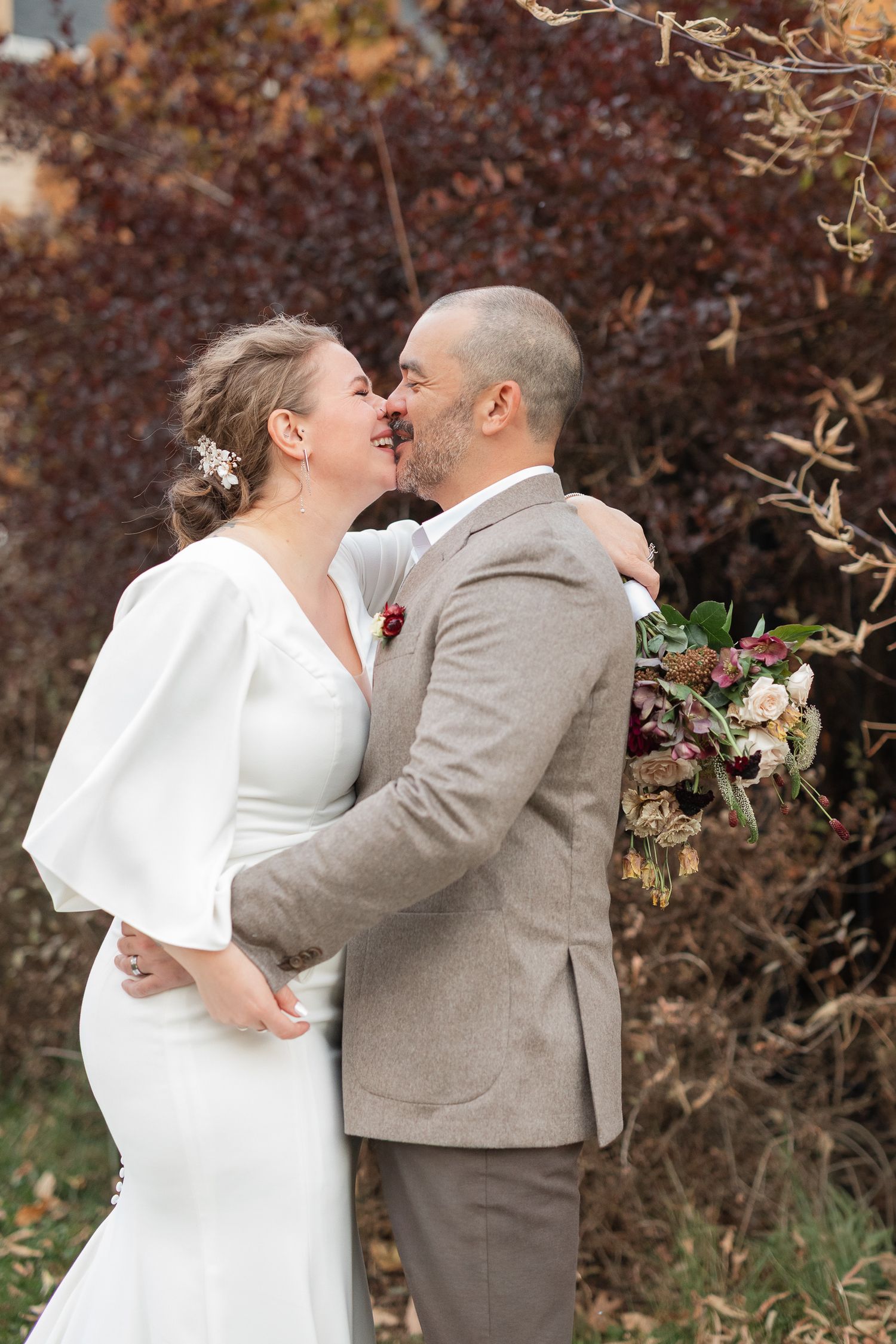A romantic wedding couple shares a tender kiss outdoors with autumn foliage in the background.