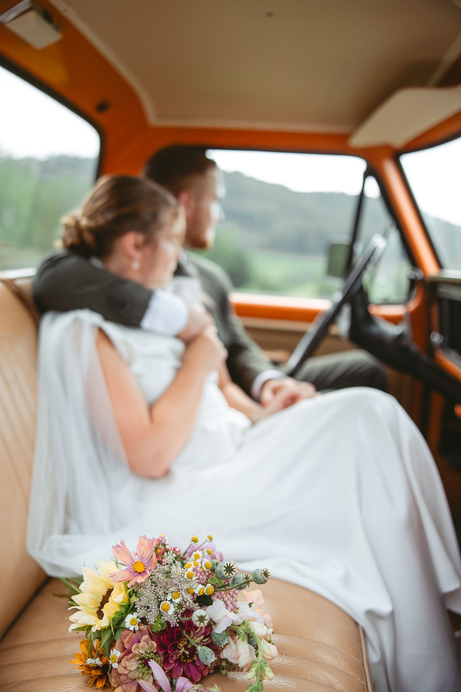 Mariée en robe blanche assise dans une voiture vintage orange, tenant un bouquet de fleurs sauvages