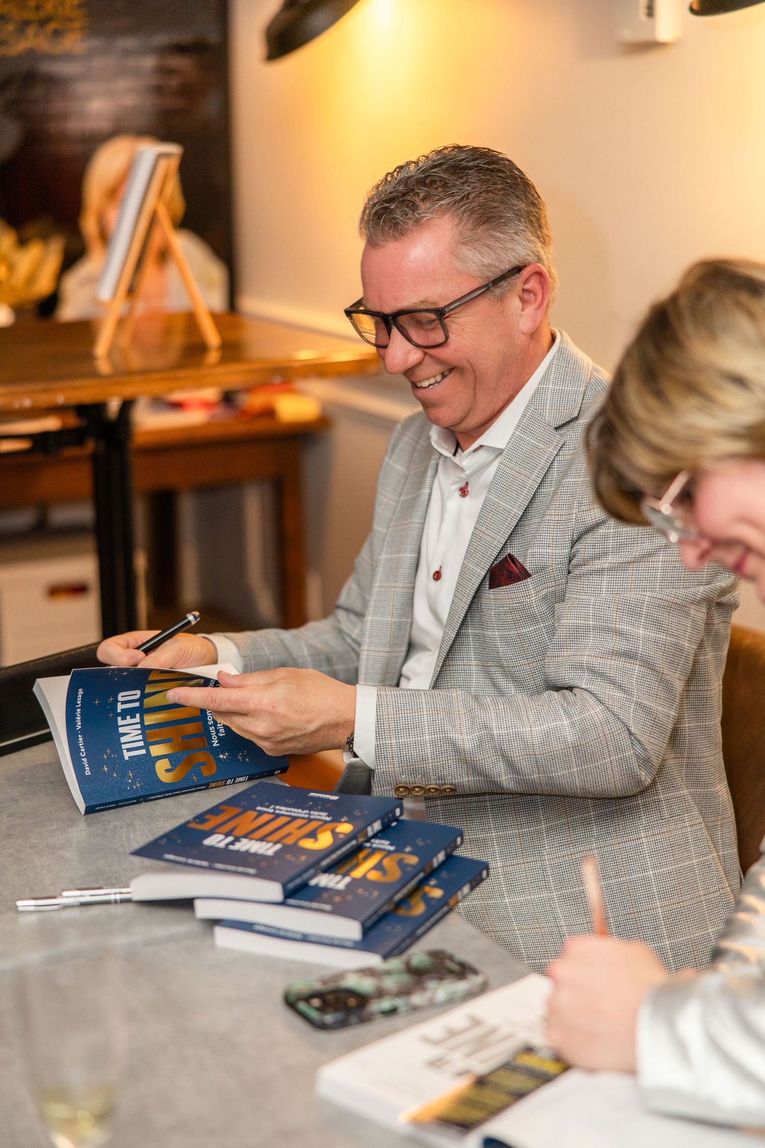 Author signing books at a table during a book signing event wearing a gray plaid blazer and glasses.