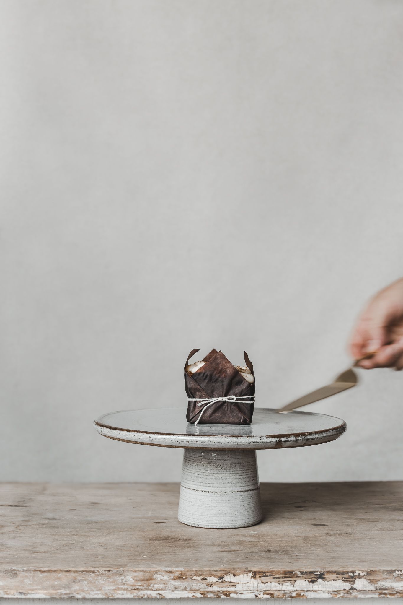 Mise en scène d’un petit gâteau sur présentoir en céramique, main en action sur fond pâle, captée pour valorisation professionnelle du produ