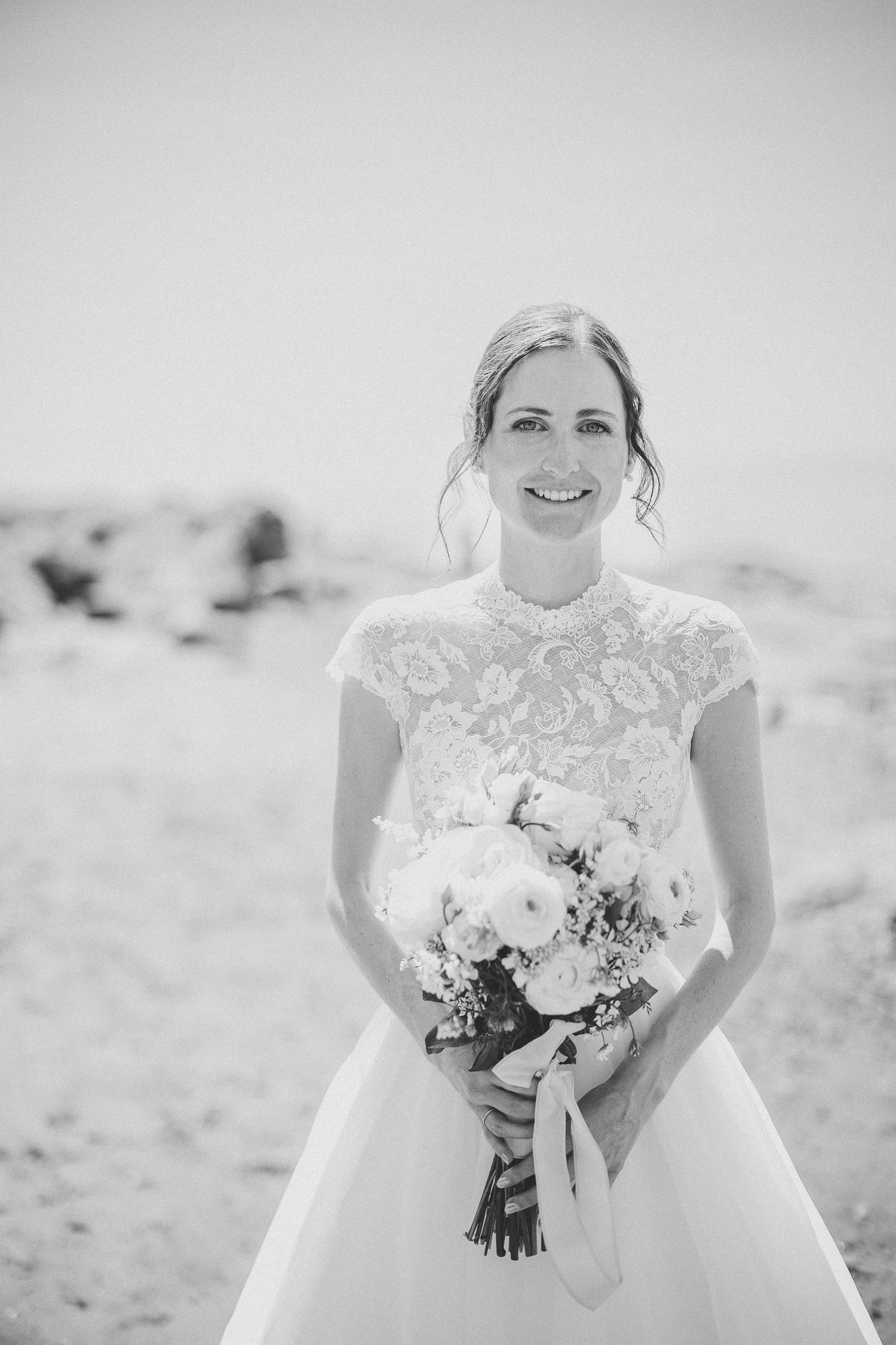 Photo noir et blanc, Mariée en robe de bal blanche tenant un bouquet floral blanc, photographiée sur une plage rocheuse lors de son mariage.