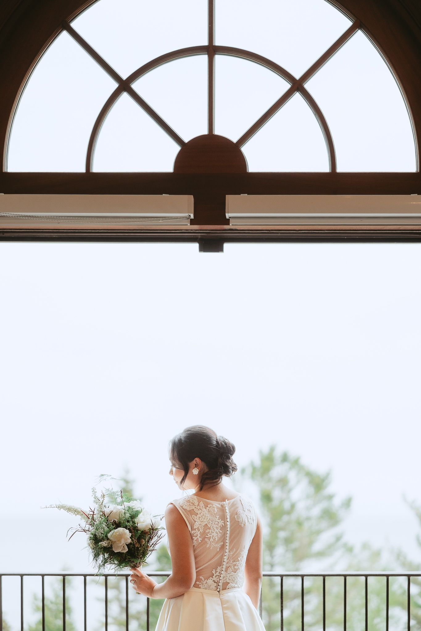 Mariée en robe de dentelle sur un balcon sous de grandes fenêtres en arche lors d’un mariage à Charlevoix.