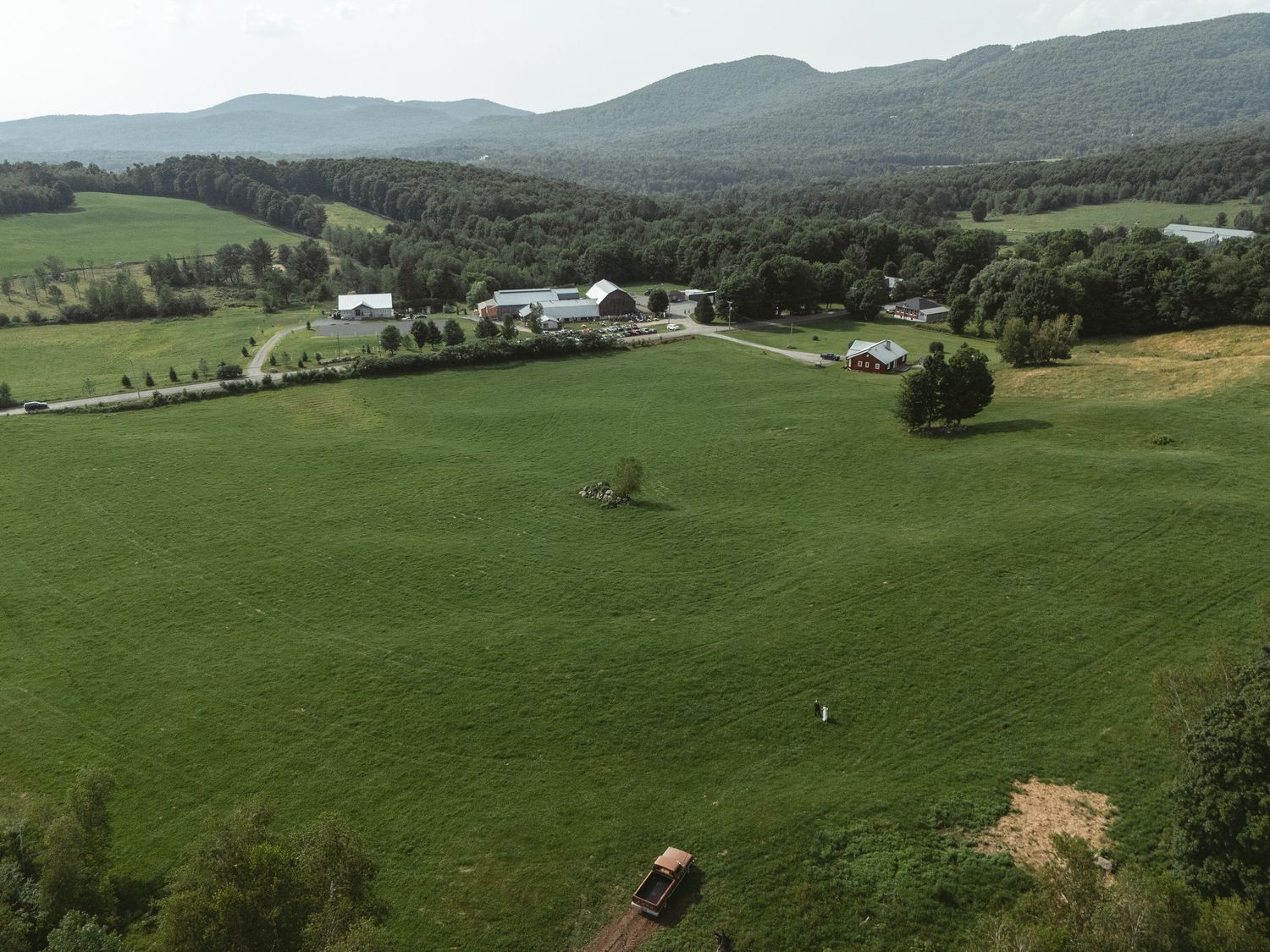 Collines verdoyantes et pâturages s’étendant dans un paysage rural avec arbres dispersés et ferme avec les mariés au loin vu de haut