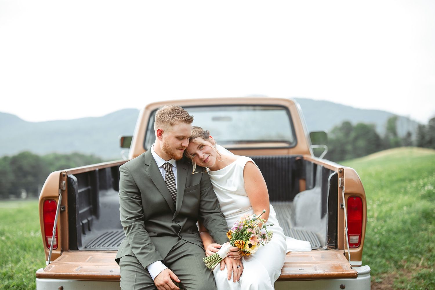 Couple en tenue de mariage assis dans la benne d’un pickup vintage, dans un champ en montagne en Estrie