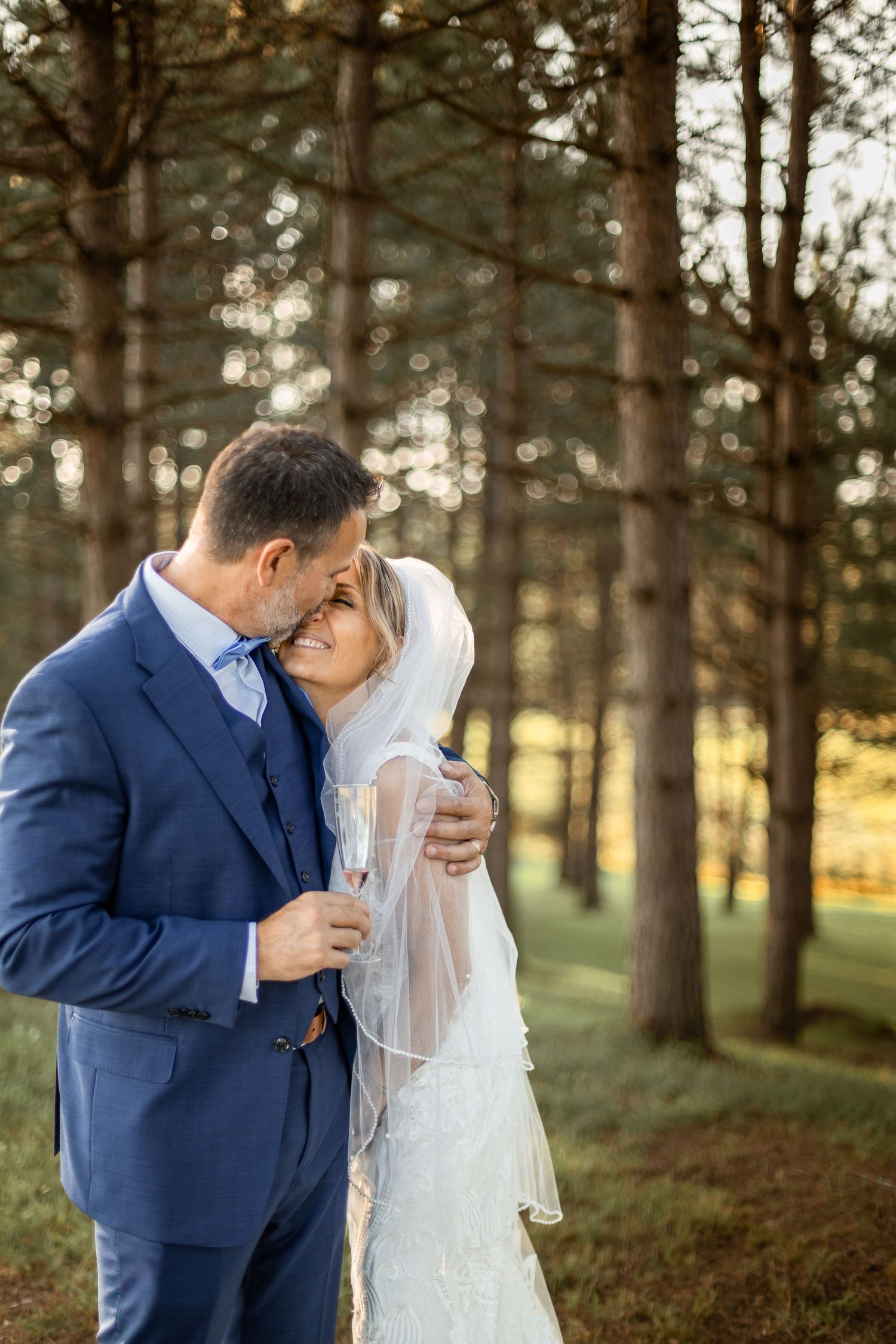 A romantic wedding couple shares tender kisses in a sunlit forest setting wearing navy blue suit and white gown.