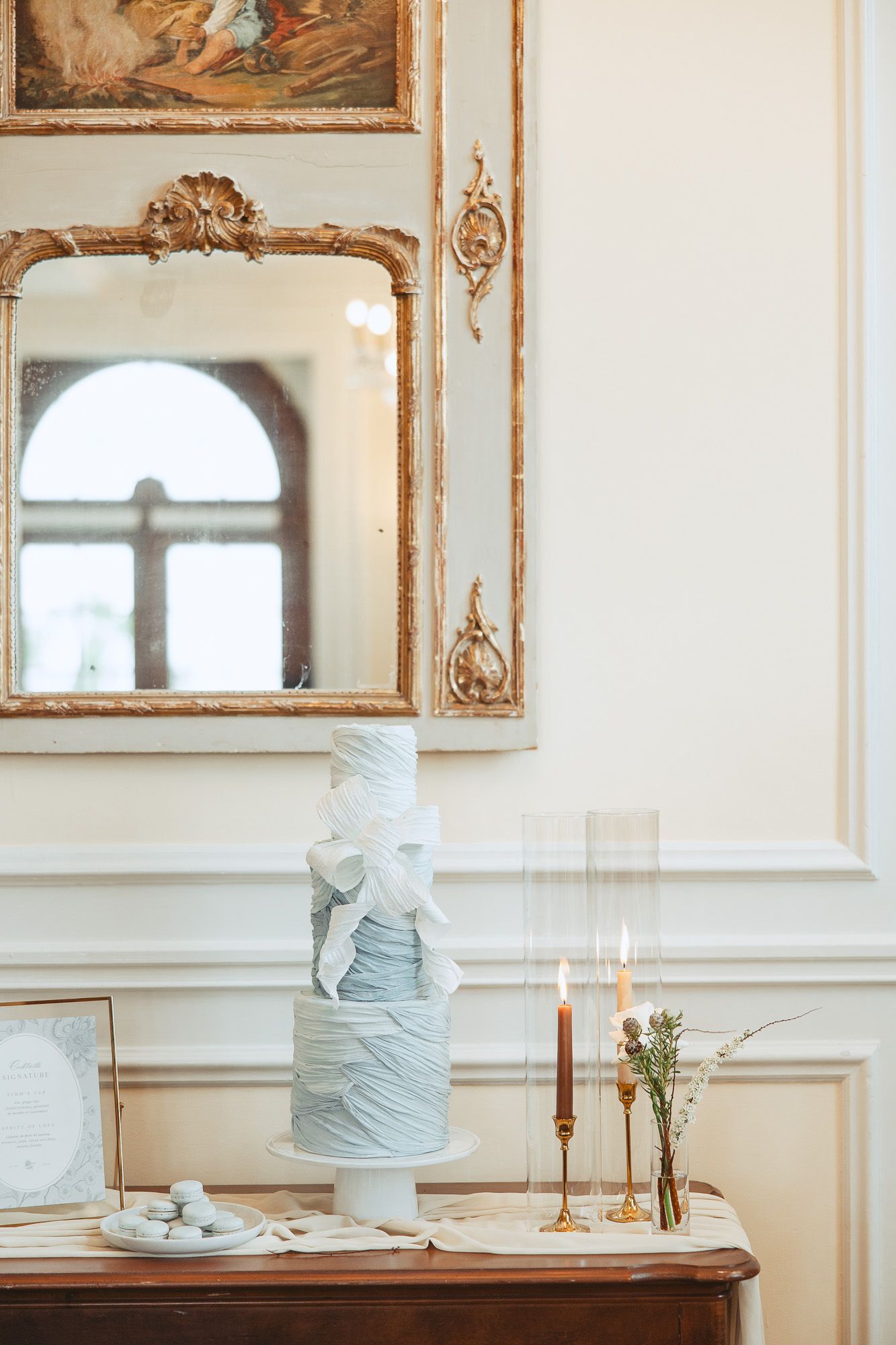 Elegant blue-gray wedding cakes displayed on wooden tables in an ornate room with gold-framed mirrors and white walls.