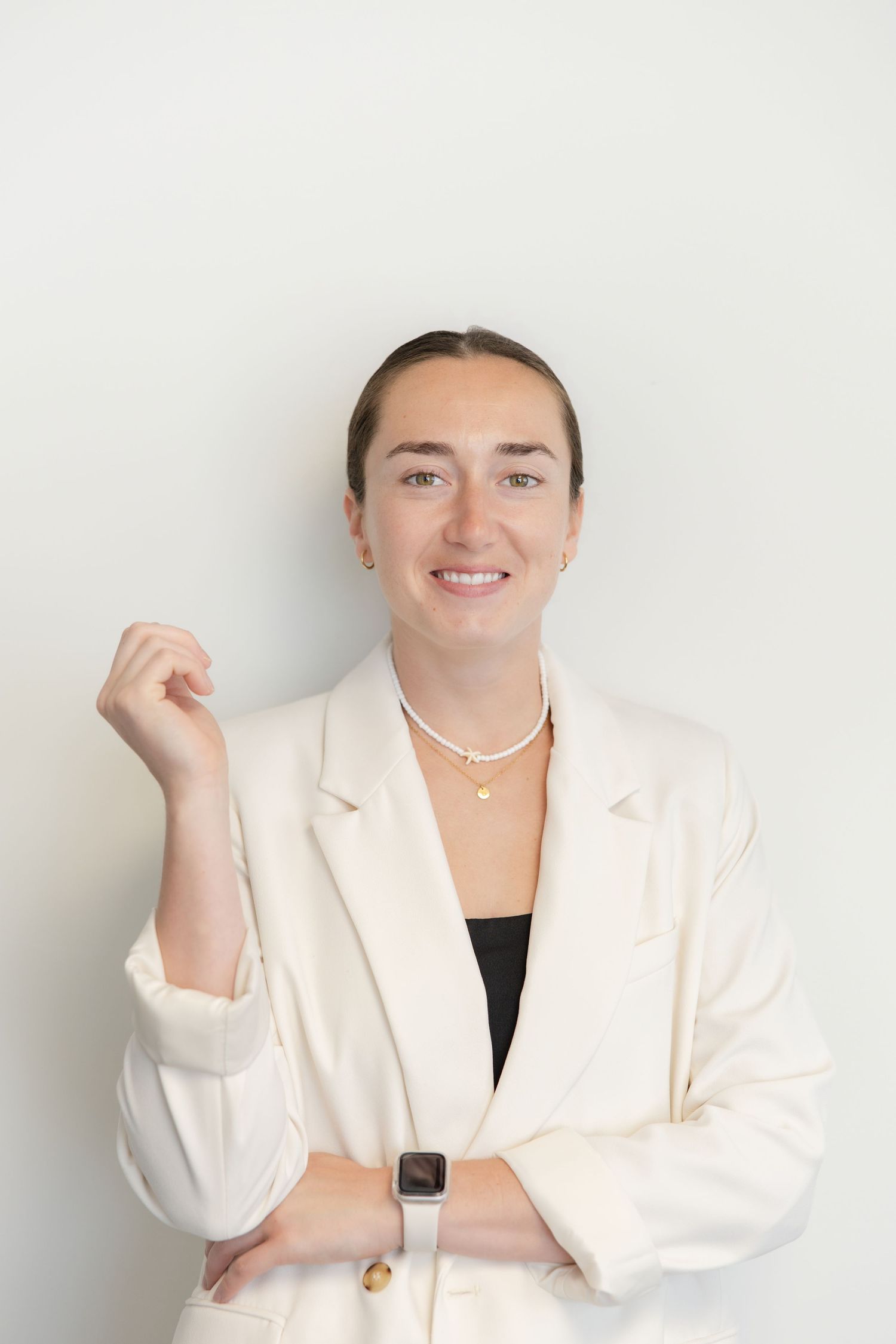 Smiling young woman in white blazer and pearl necklace with smartwatch poses confidently against light background.