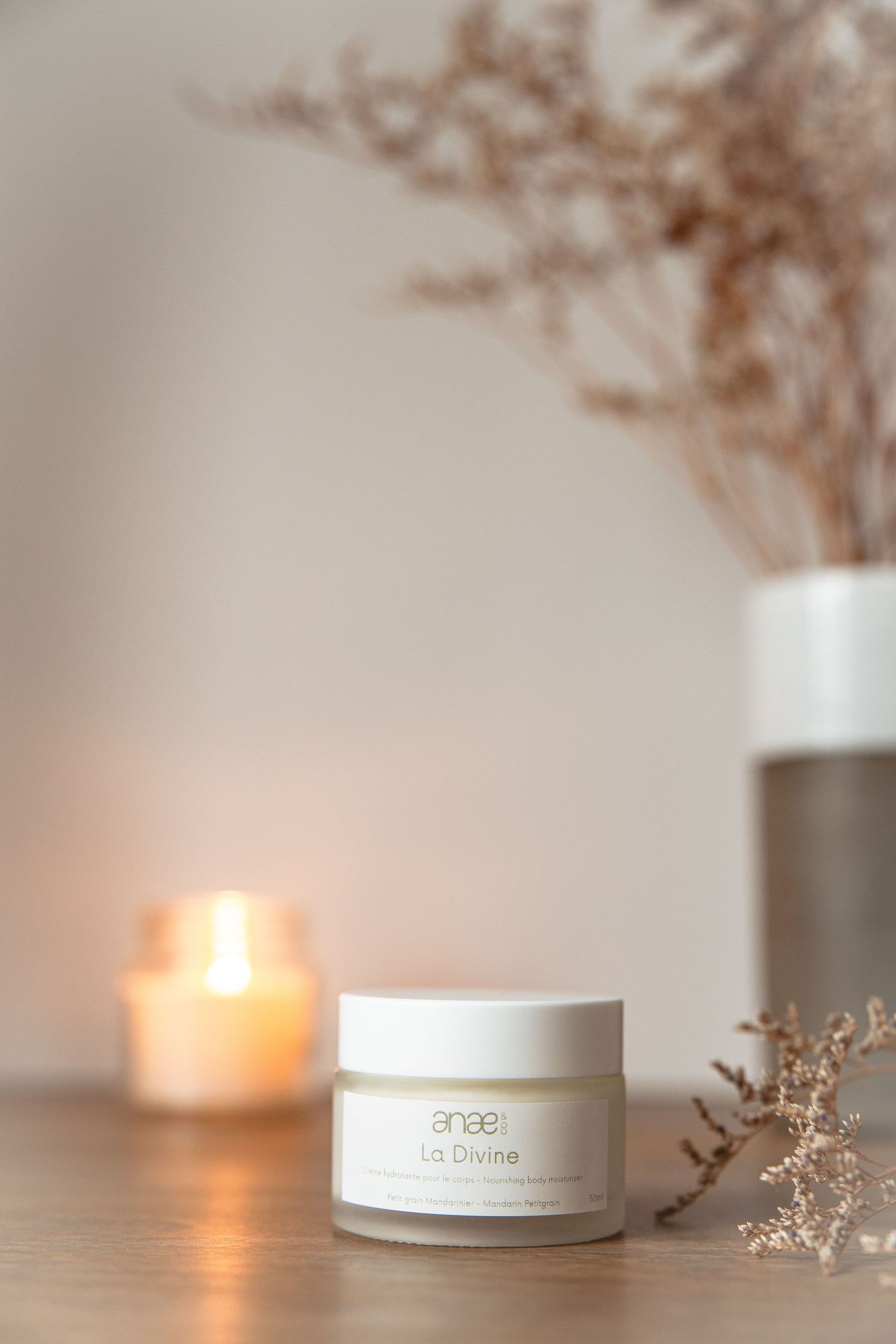 Minimalist beauty product jars with dried flowers arranged on white surface in warm candlelit atmosphere.