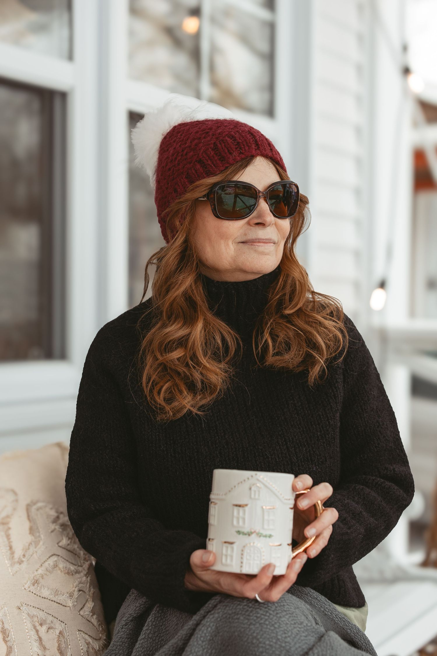 Person in black sweater and burgundy beanie holds white gift box while sitting by window in winter setting.
