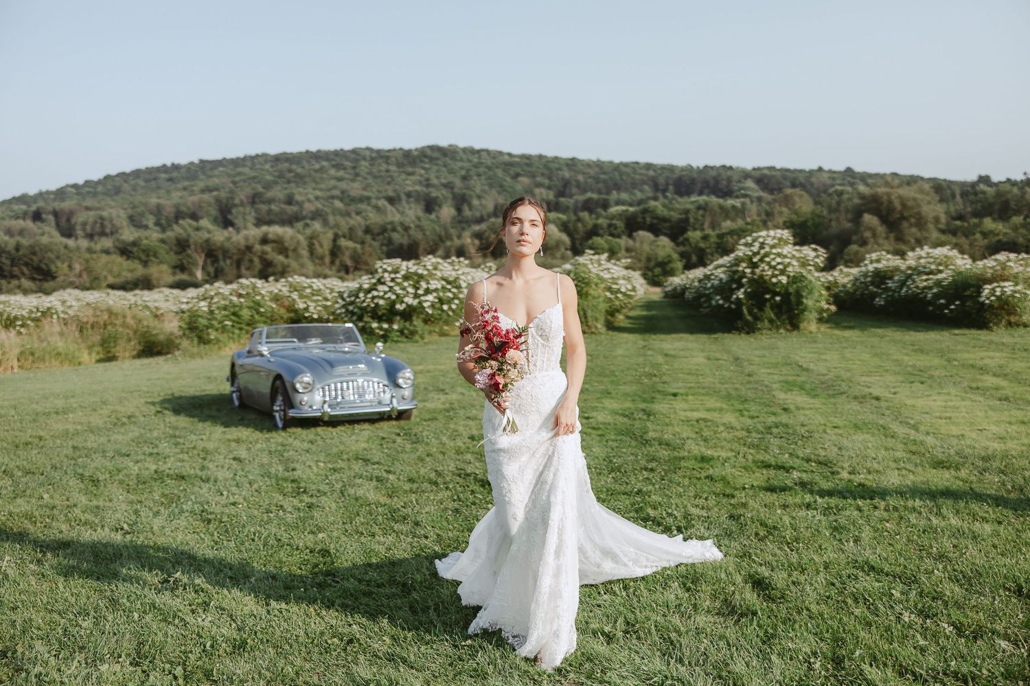 Mariée qui marche bouquet à la main avec un champ de sureau et montagne en arrière plan et une voiture décapotable bleu vintage