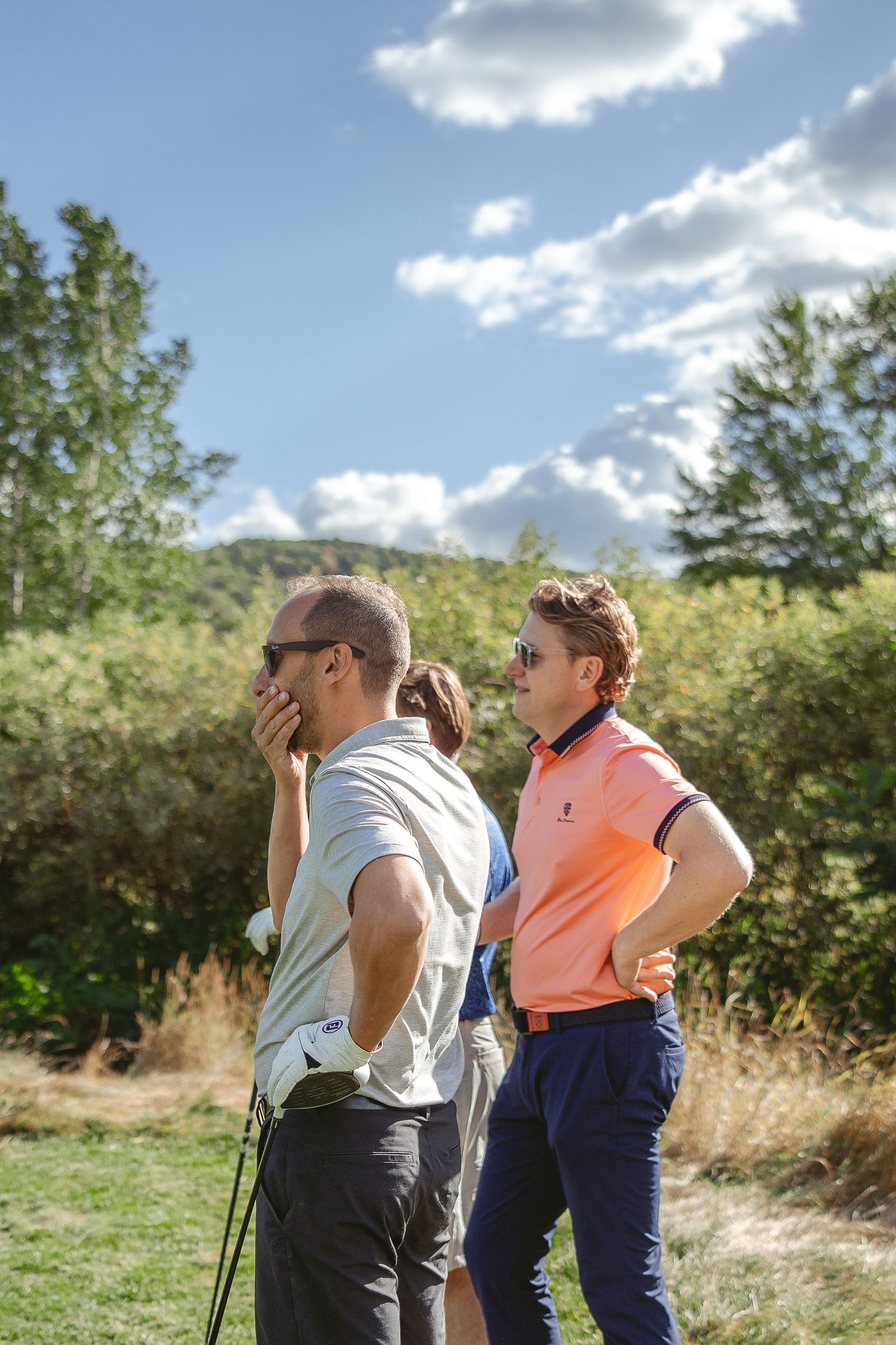 Two people use binoculars to observe wildlife while standing in a sunny meadow on a partly cloudy day.