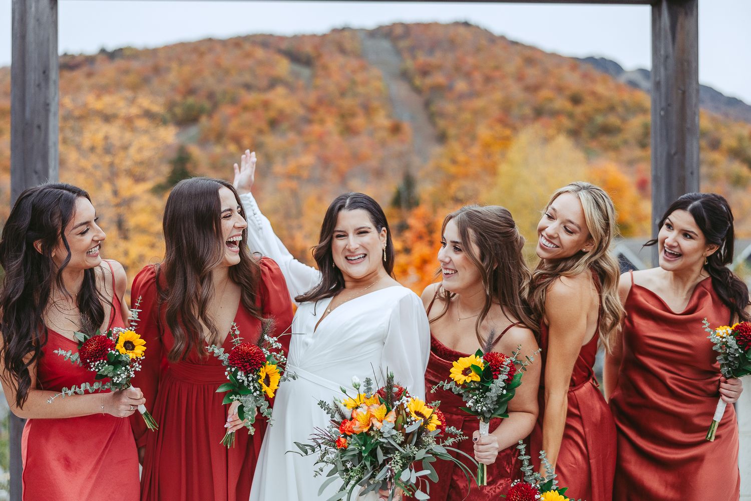 Le cortège de mariage, vêtu de robes bourgogne, rit ensemble lors d’une séance photo automnale, avec en arrière-plan un feuillage d'automne