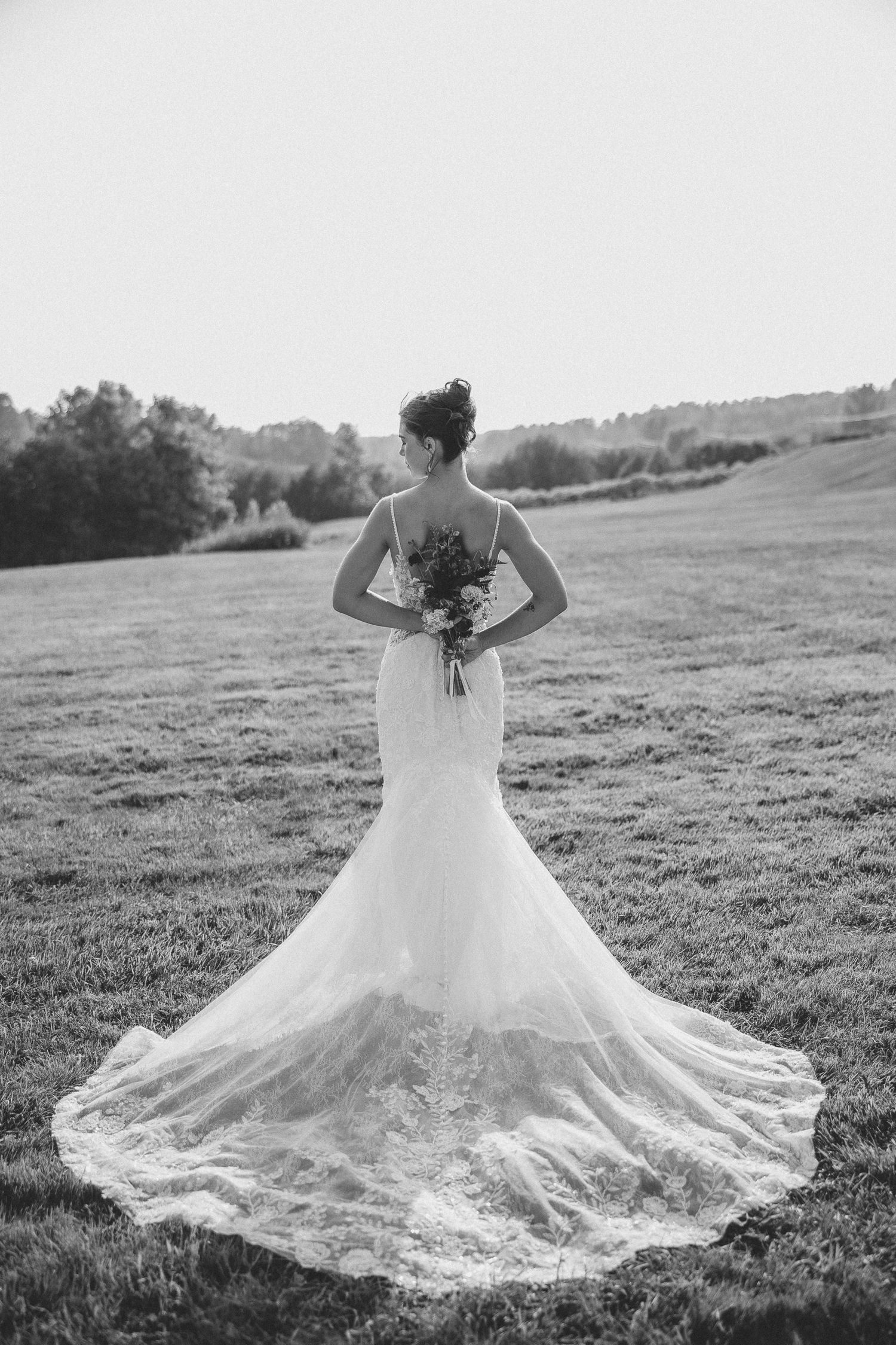 Photo de mariage en noir et blanc montrant une mariée en robe de dentelle fluide à longue traîne, debout dans un champ au coucher du soleil.